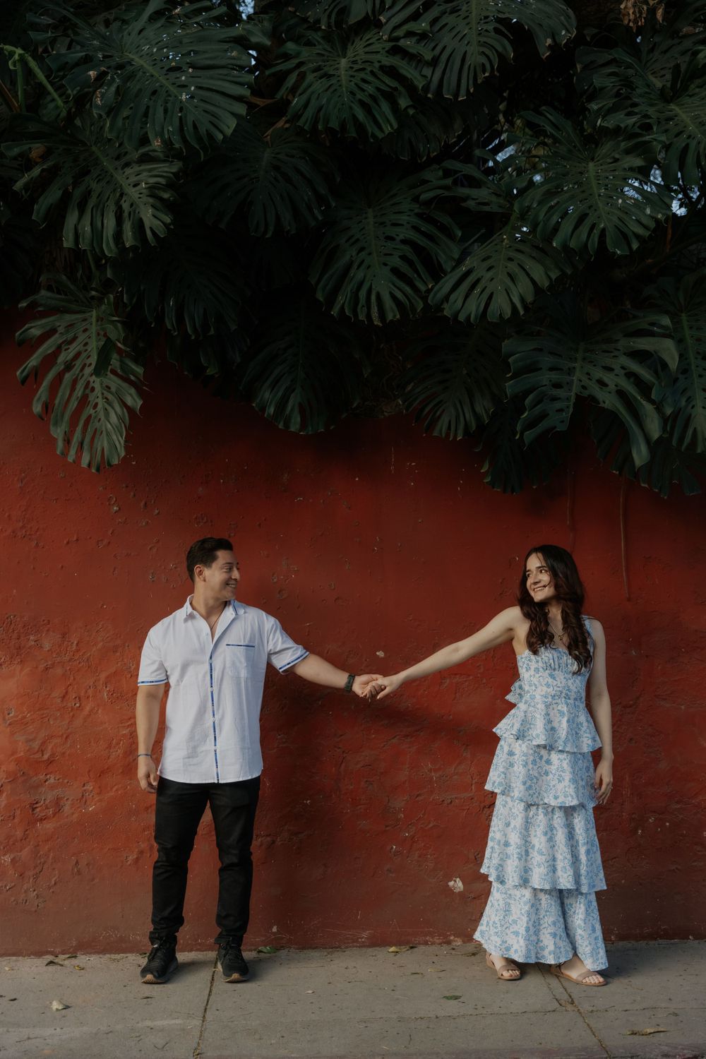 A couple in light summer clothing poses against a red wall with tropical monstera leaves overhead.