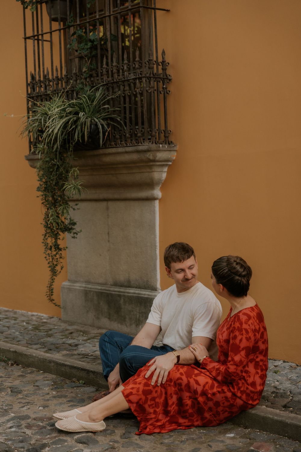 A couple sitting casually on a cobblestone street against an orange wall with decorative ironwork and plants.