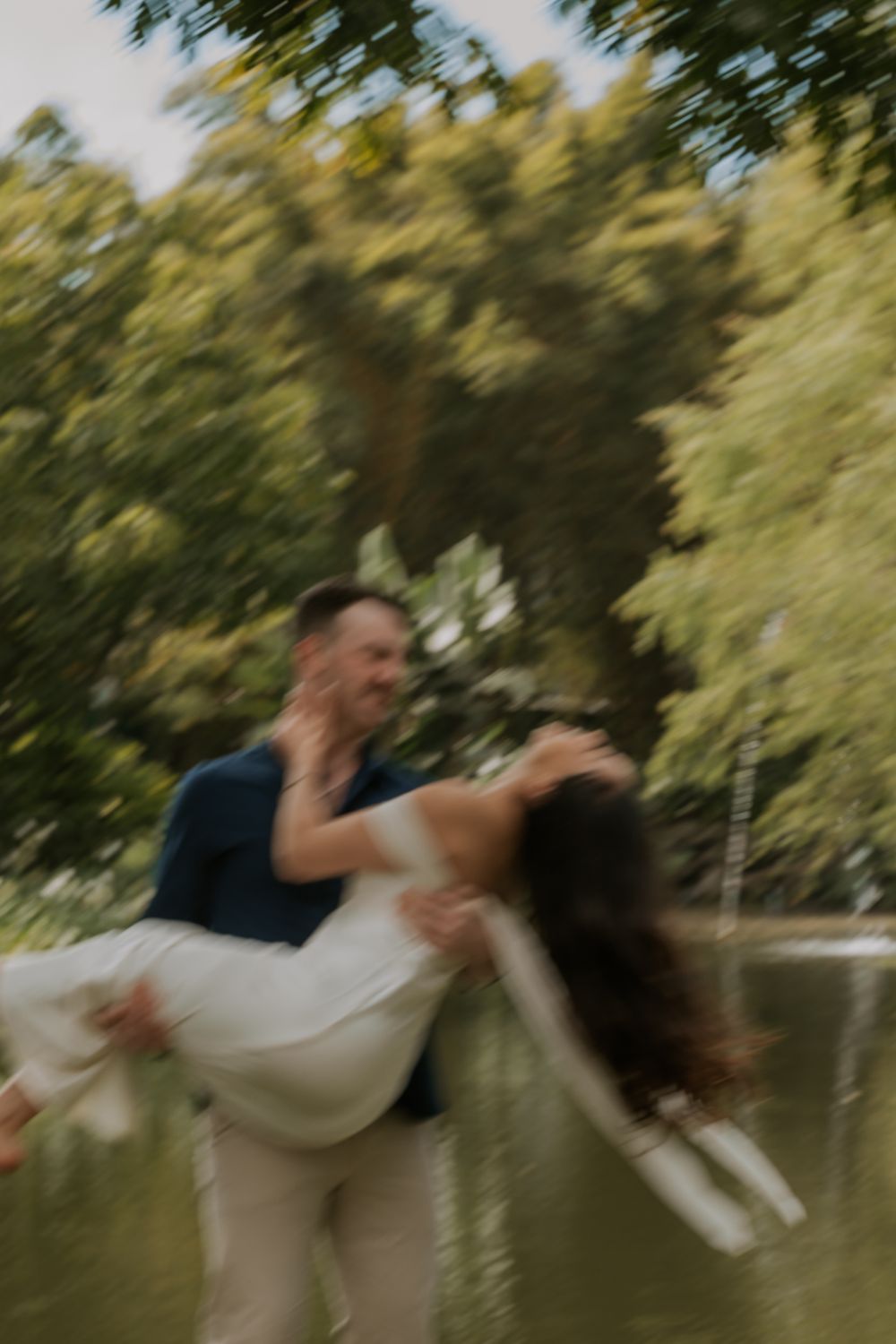 A romantic couple shares a playful dip pose in a garden setting with blurred trees in the background.