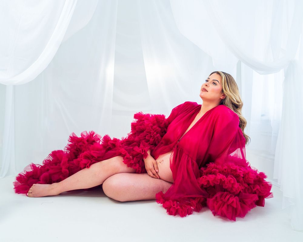 Model in flowing red dress reclines on white studio floor. Maternity Photos.