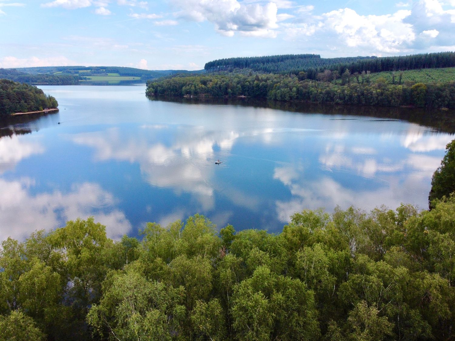 Tranquil lake surrounded by forested cliffs reflects puffy white clouds in blue sky on a sunny summer day.
