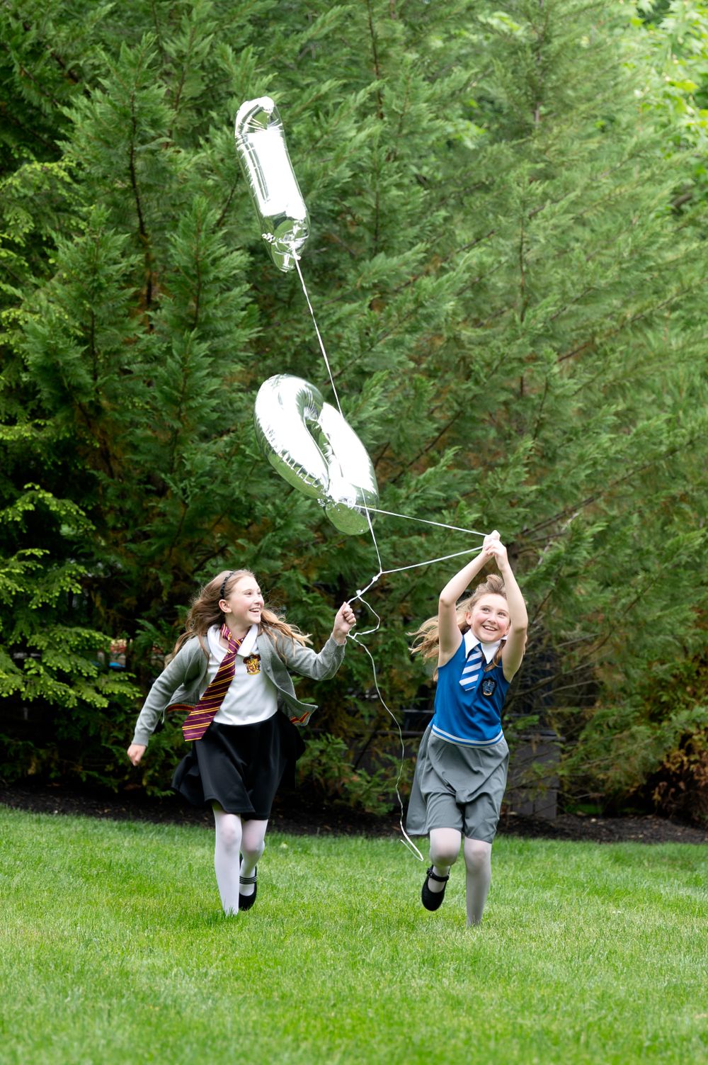 Two children running on grass with white balloons attached to strings on a sunny day.