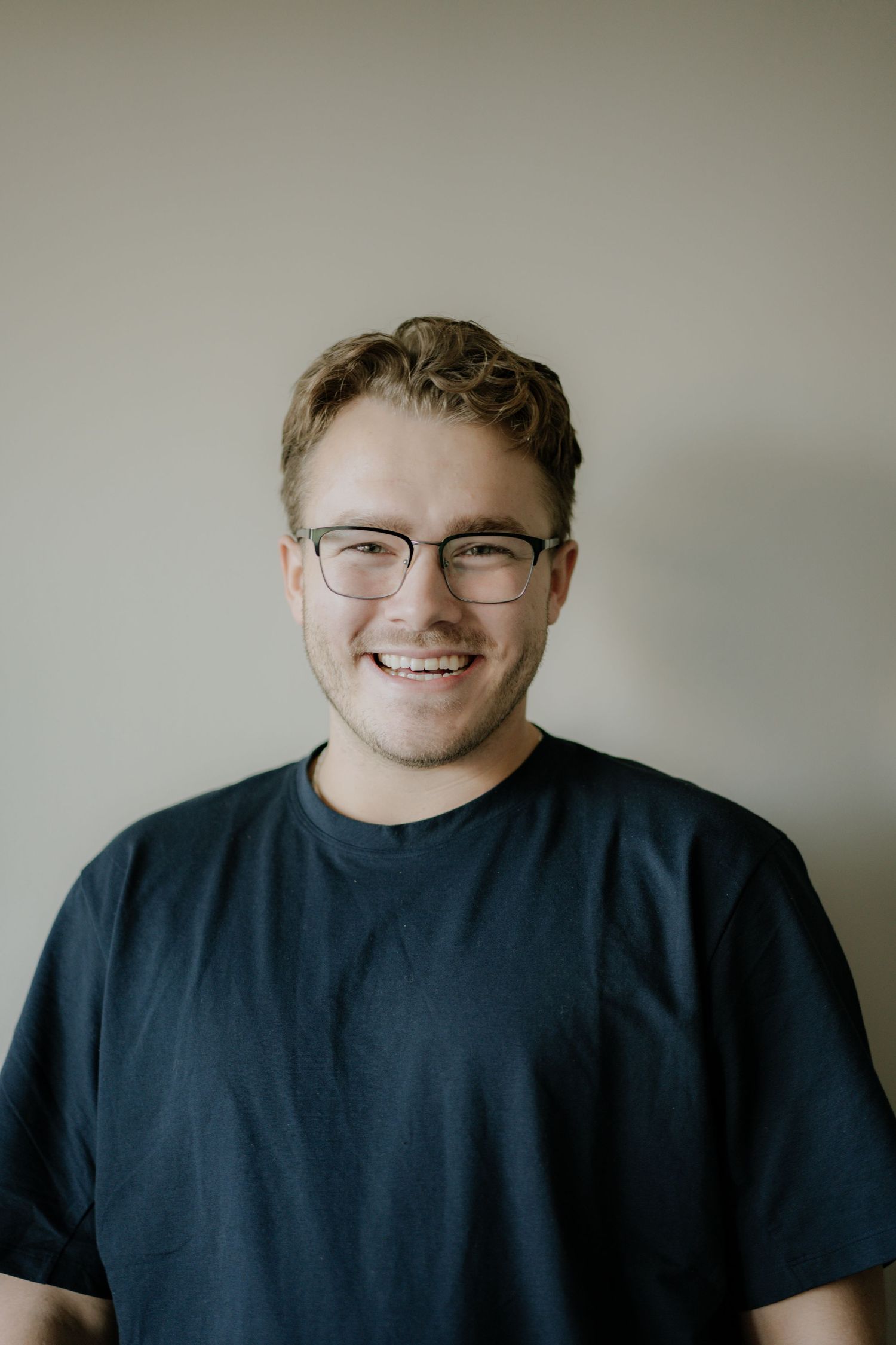 Smiling young man with glasses wearing a dark navy t-shirt posing against a neutral wall.