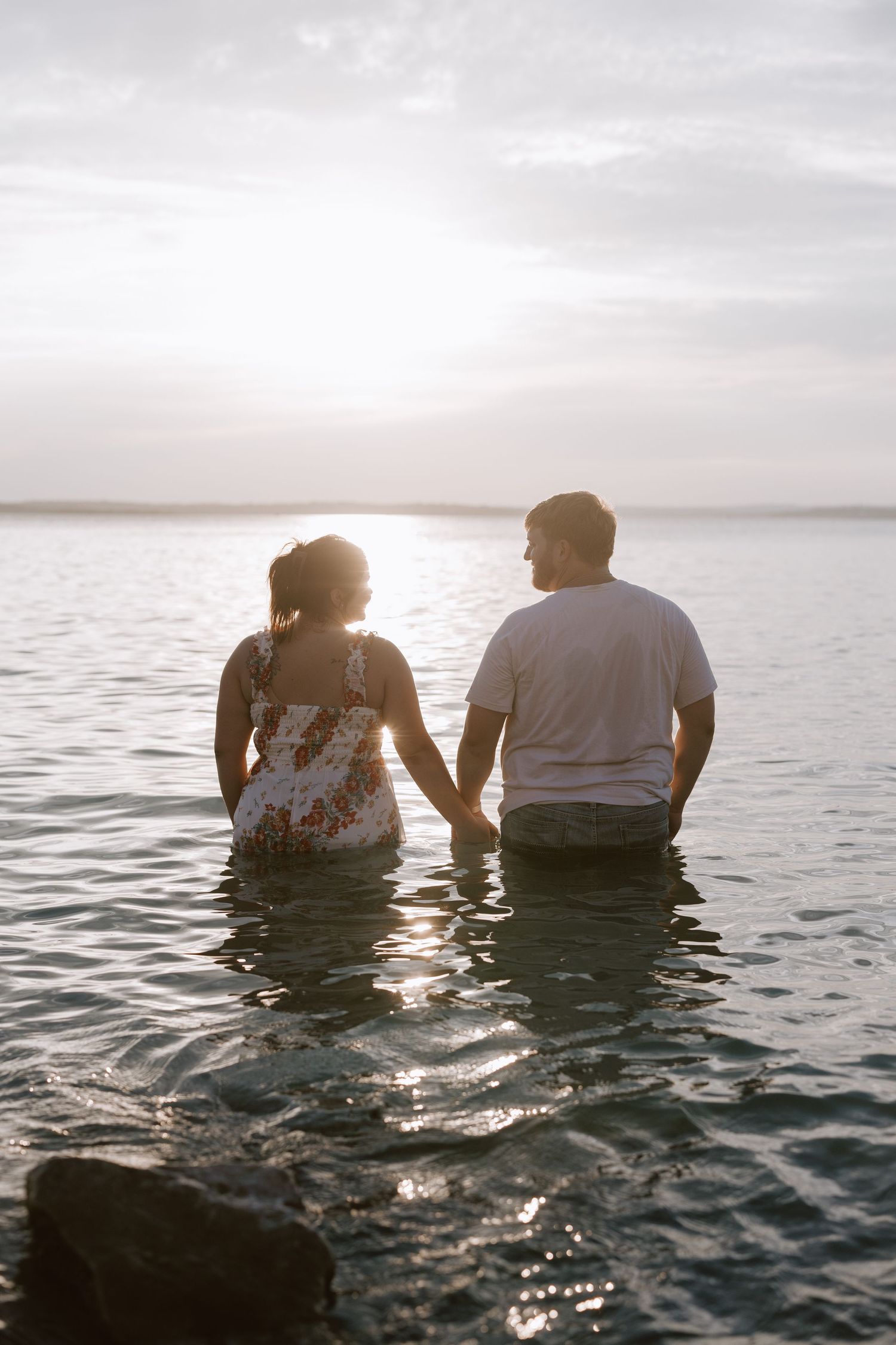 A couple sits together in shallow water during sunset, creating a romantic silhouette against the shimmering lake.