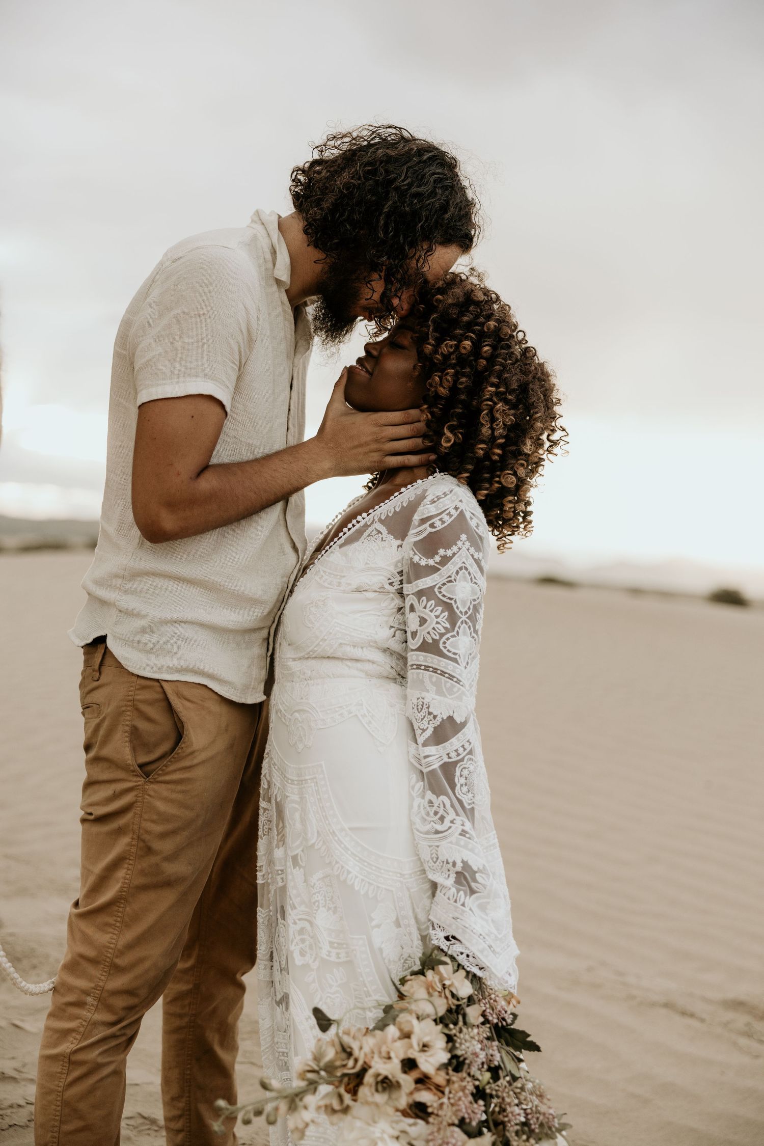 Intimate wedding portrait of couple embracing on sandy beach with bouquet.