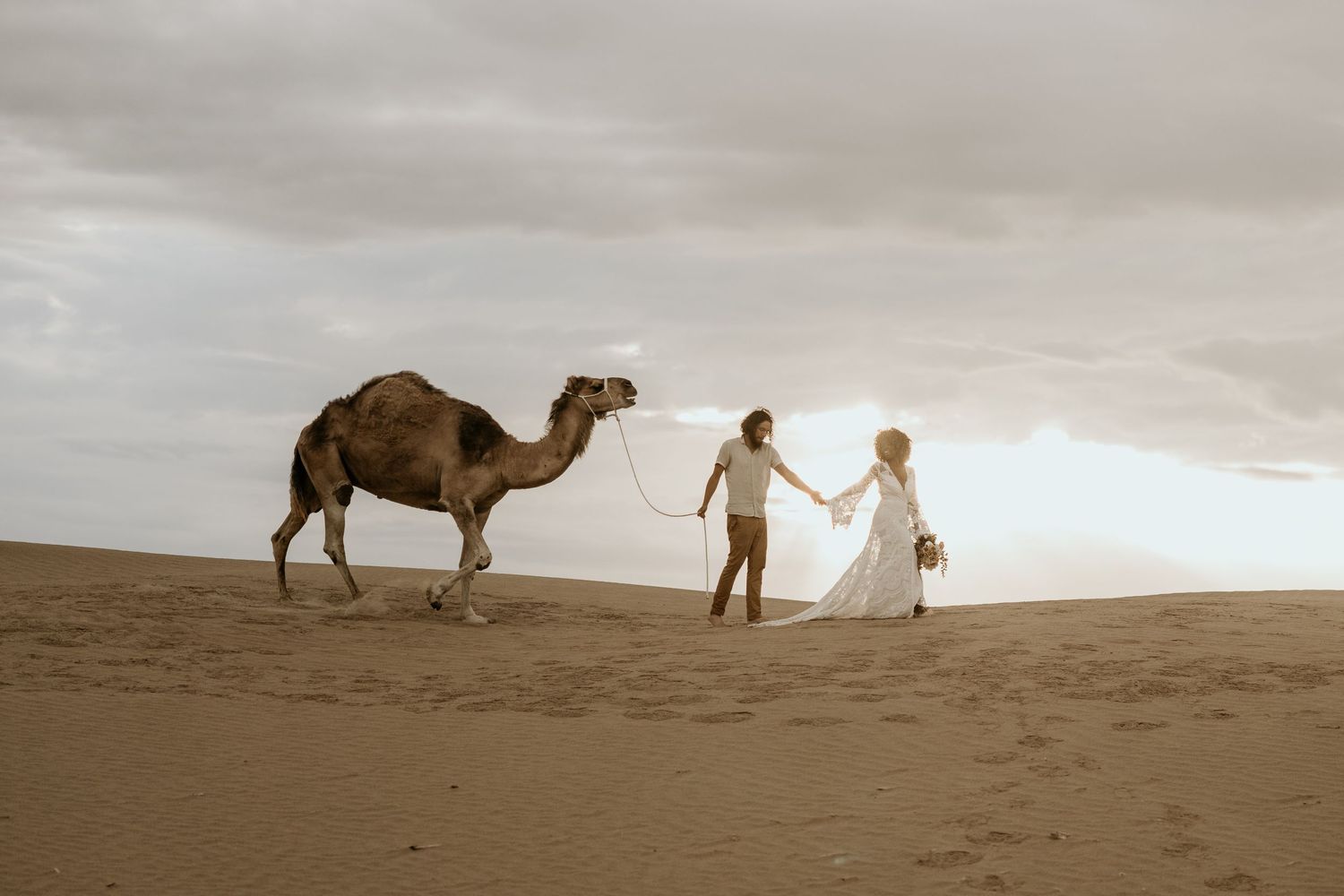 A serene desert wedding scene with a couple walking alongside a camel in sepia tones.
