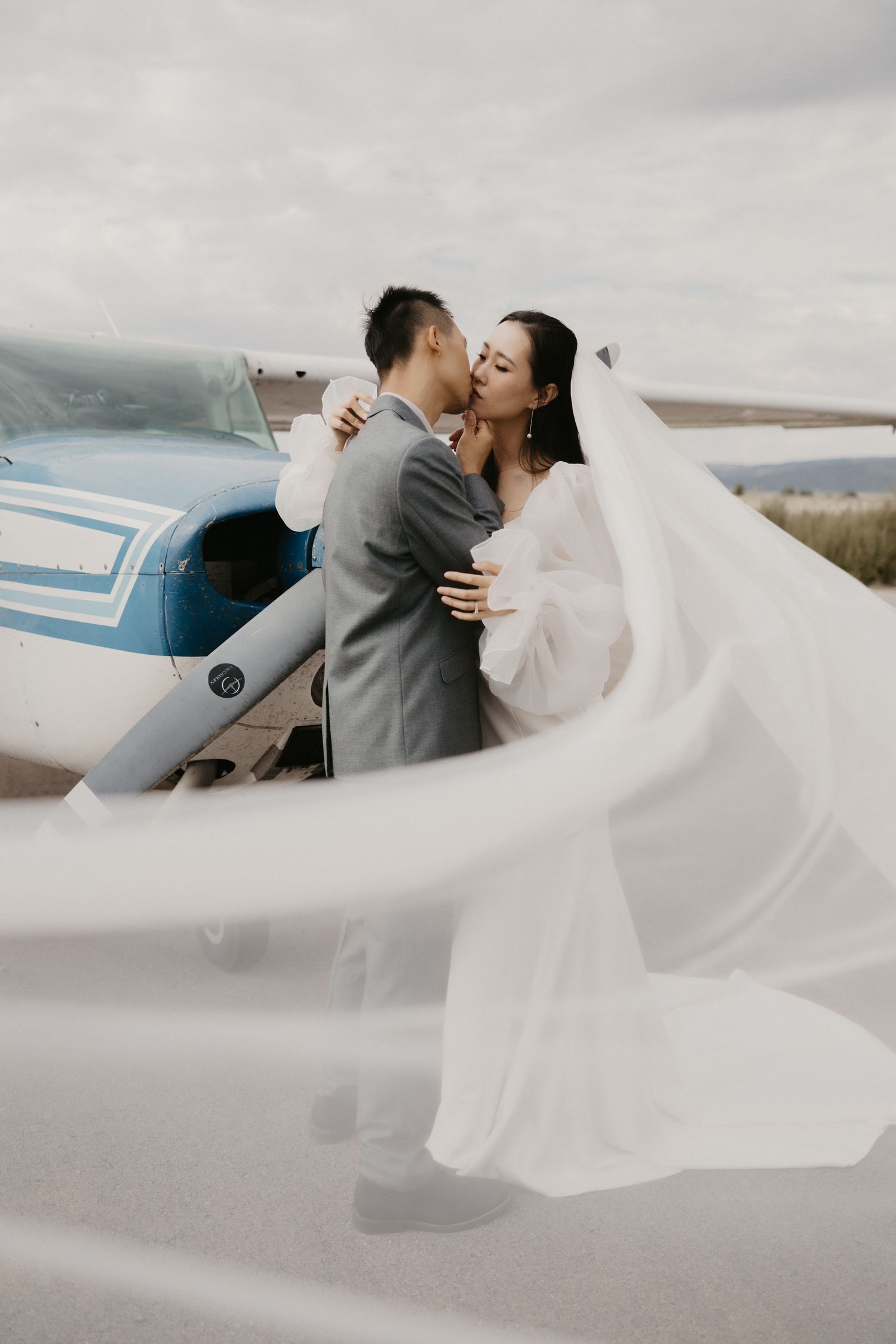 Bride and groom sharing romantic kiss by small airplane with flowing veil.