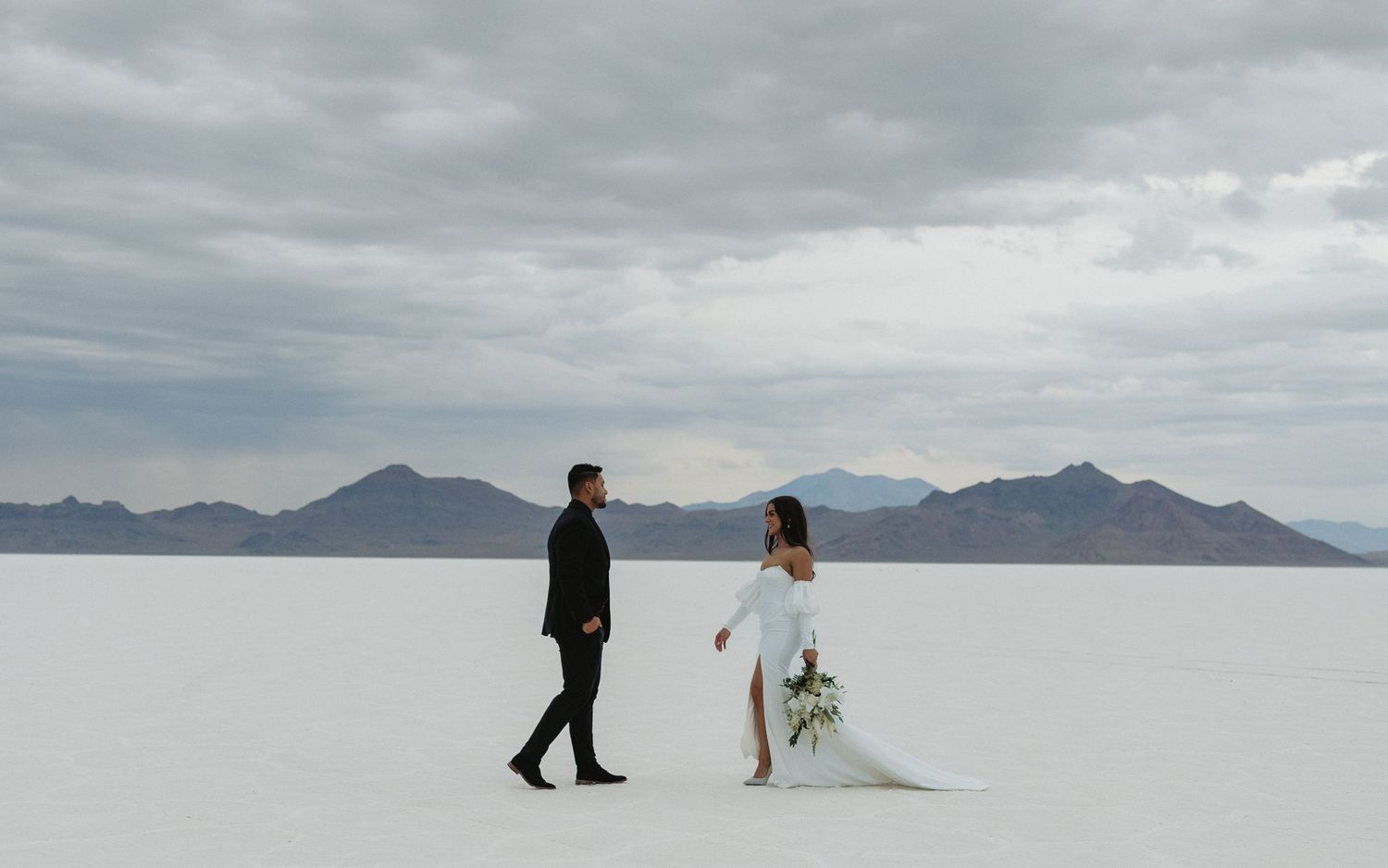 A couple in wedding attire walking on the dramatic salt flats with mountains in the distance.