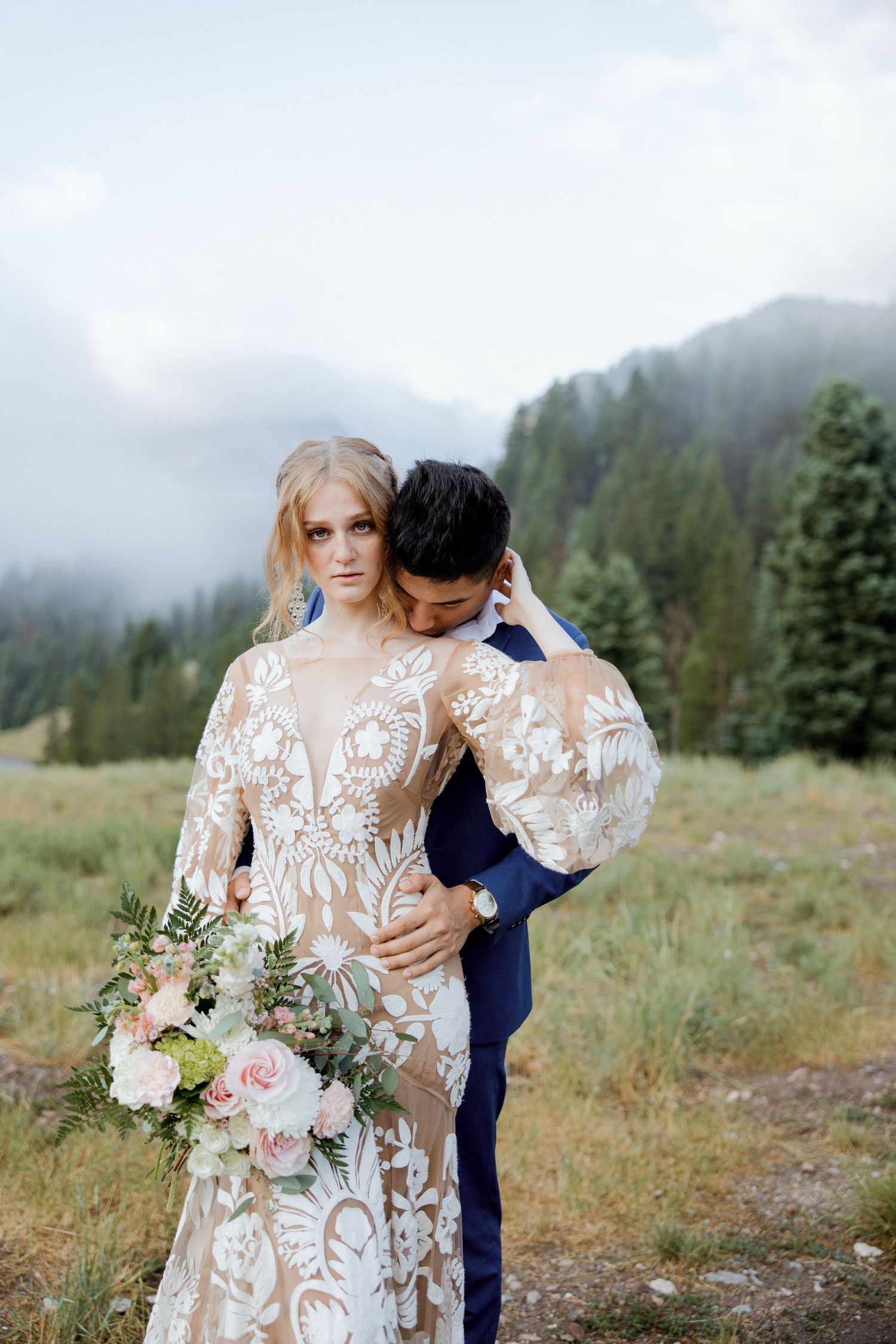 Romantic wedding couple embracing in mountain meadow with lace dress and florals.