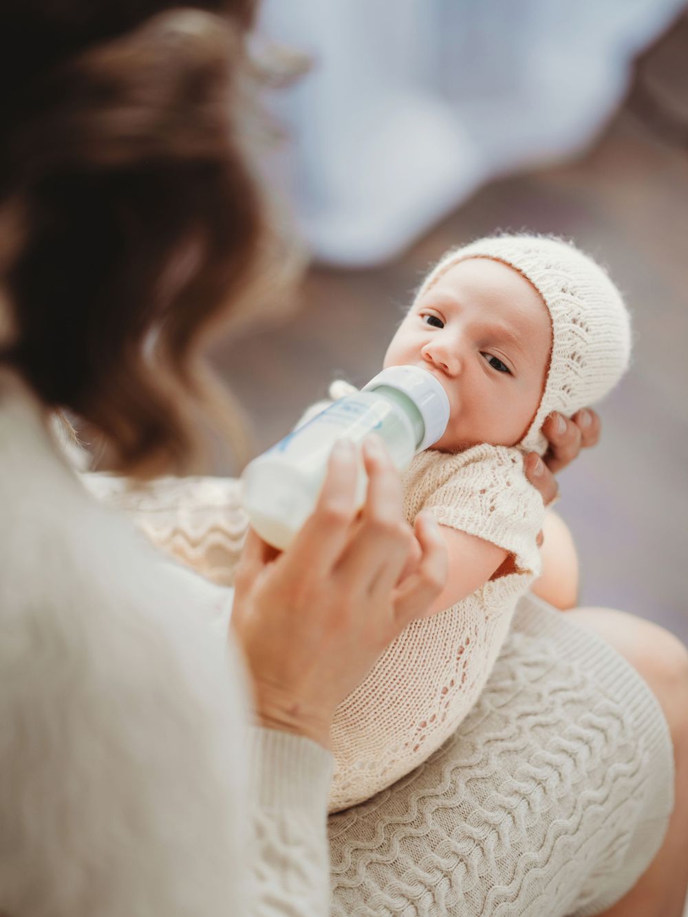 Baby wearing a white knit outfit and bonnet being bottle fed while sitting on a cozy knitted blanket in Bellevue studio.