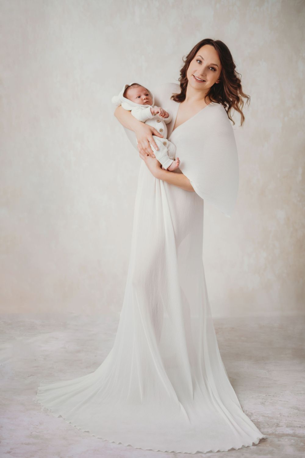 A woman in a flowing white gown holds a sleeping newborn baby in a soft, ethereal studio portrait setting in Bellevue studio.