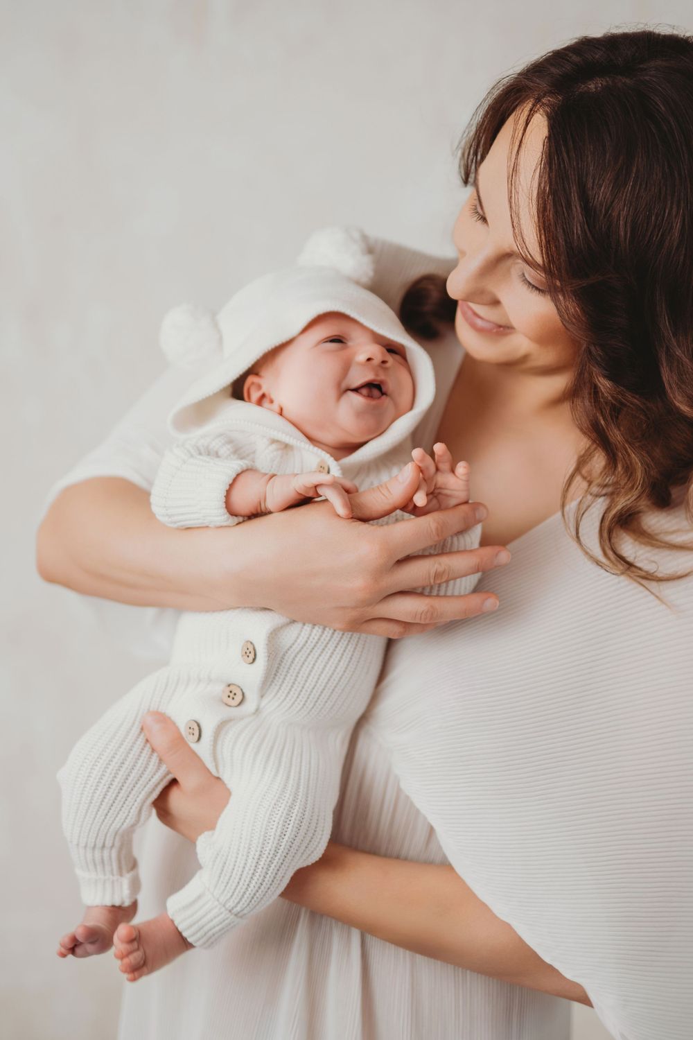 An intimate moment shows a mother in white holding a laughing baby wearing a cozy knit outfit and bear ear hat in Bellevue studio.