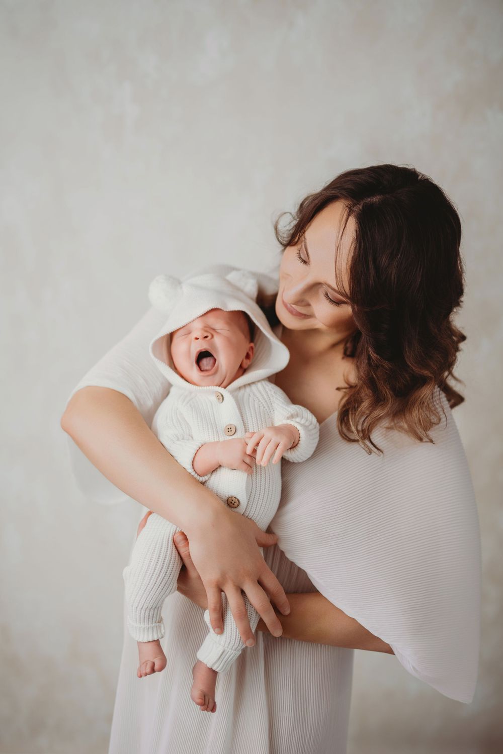 A tender moment between a parent and yawning newborn baby dressed in white against a neutral background in Bellevue studio.