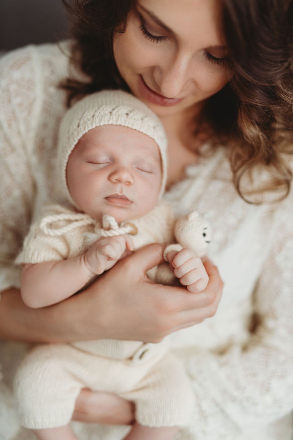 A sleeping newborn baby wearing a white knit outfit and hat is held tenderly while clutching a small stuffed toy in Bellevue studio.