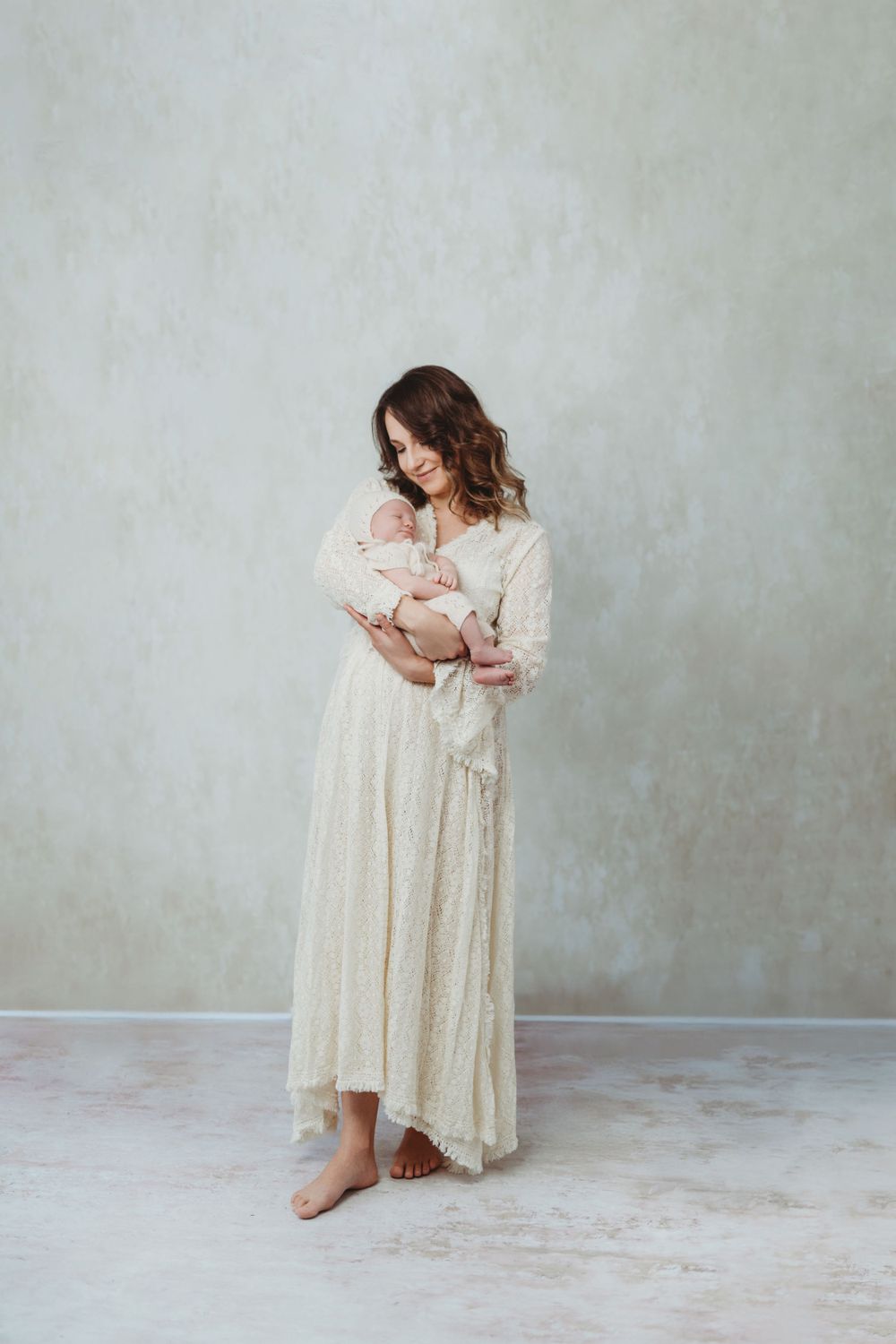 A tender motherhood portrait with a woman in a flowing cream lace dress holding a newborn baby against a light gray backdrop in Bellevue.
