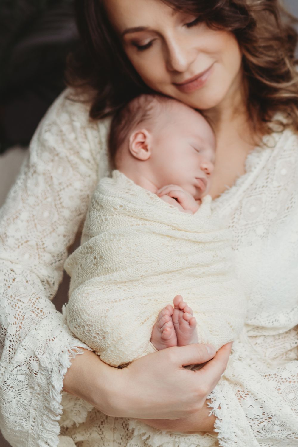 Tender moment between mother and sleeping newborn baby wrapped in white lace and knit blankets in Bellevue studio.