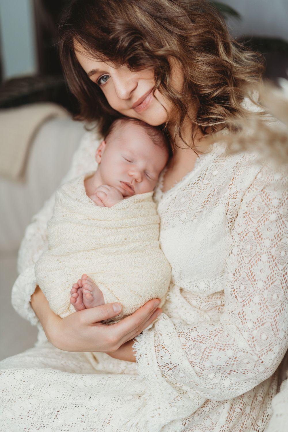 A sleeping newborn baby wrapped in white swaddle cloth while being held in white lace dress in Bellevue studio.