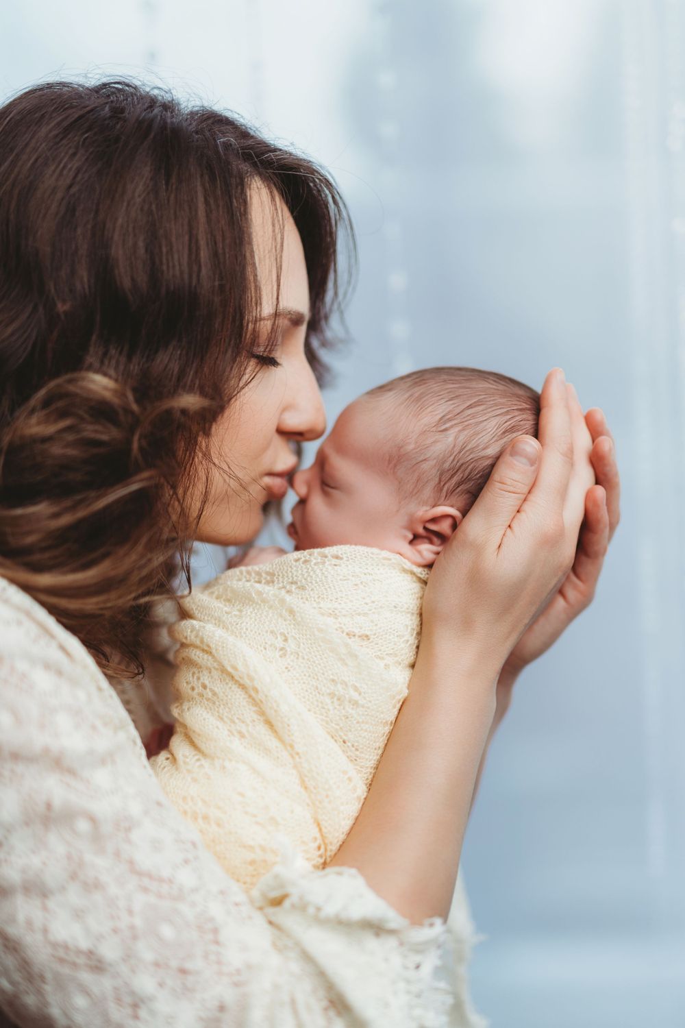Tender moment between mother and newborn baby wrapped in cream blanket against soft blue background in Bellevue studio.