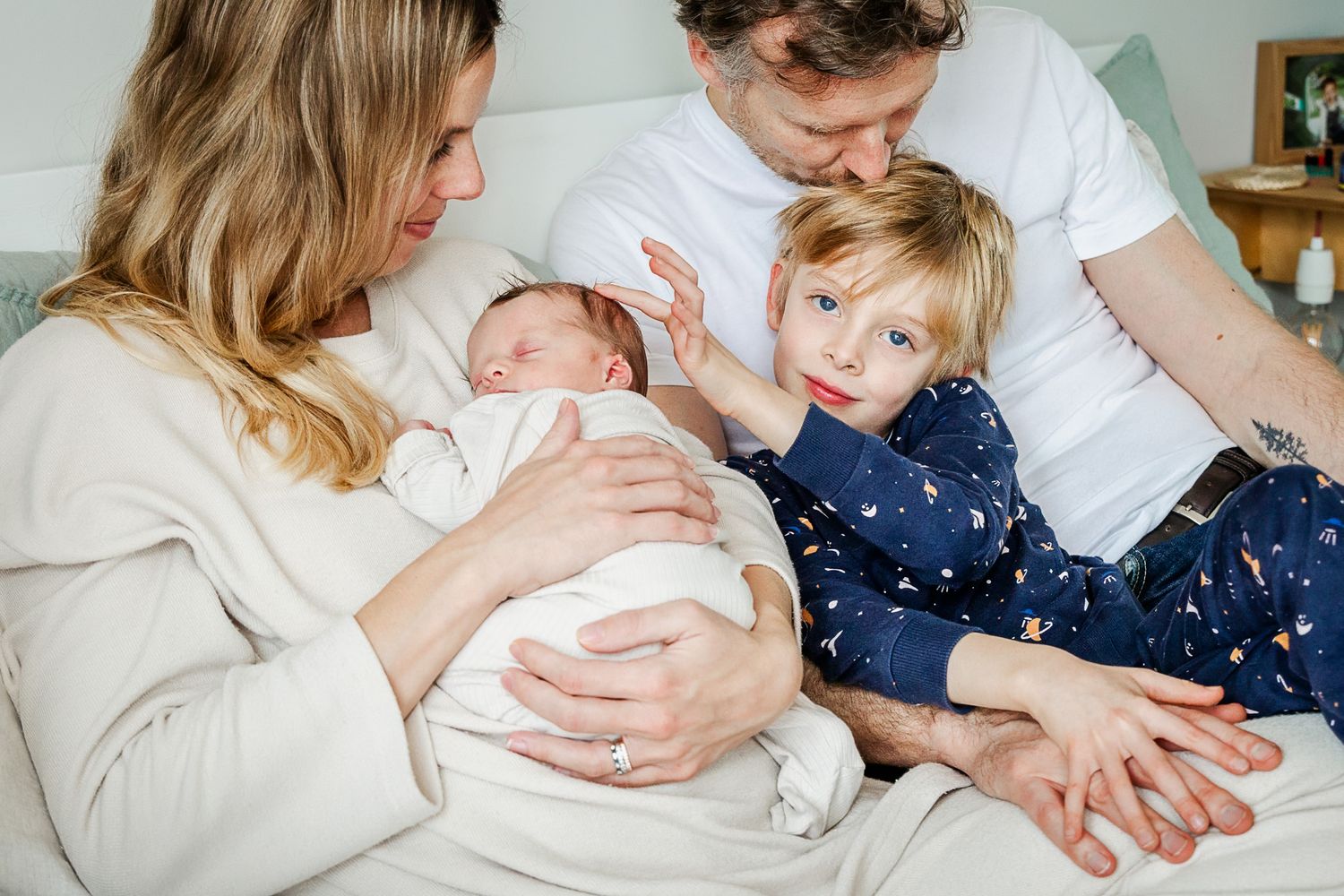A family cuddle on the bd at home. Mum holds her newborn baby as dad kisses their older son on the head. Natural Bristol family photographer