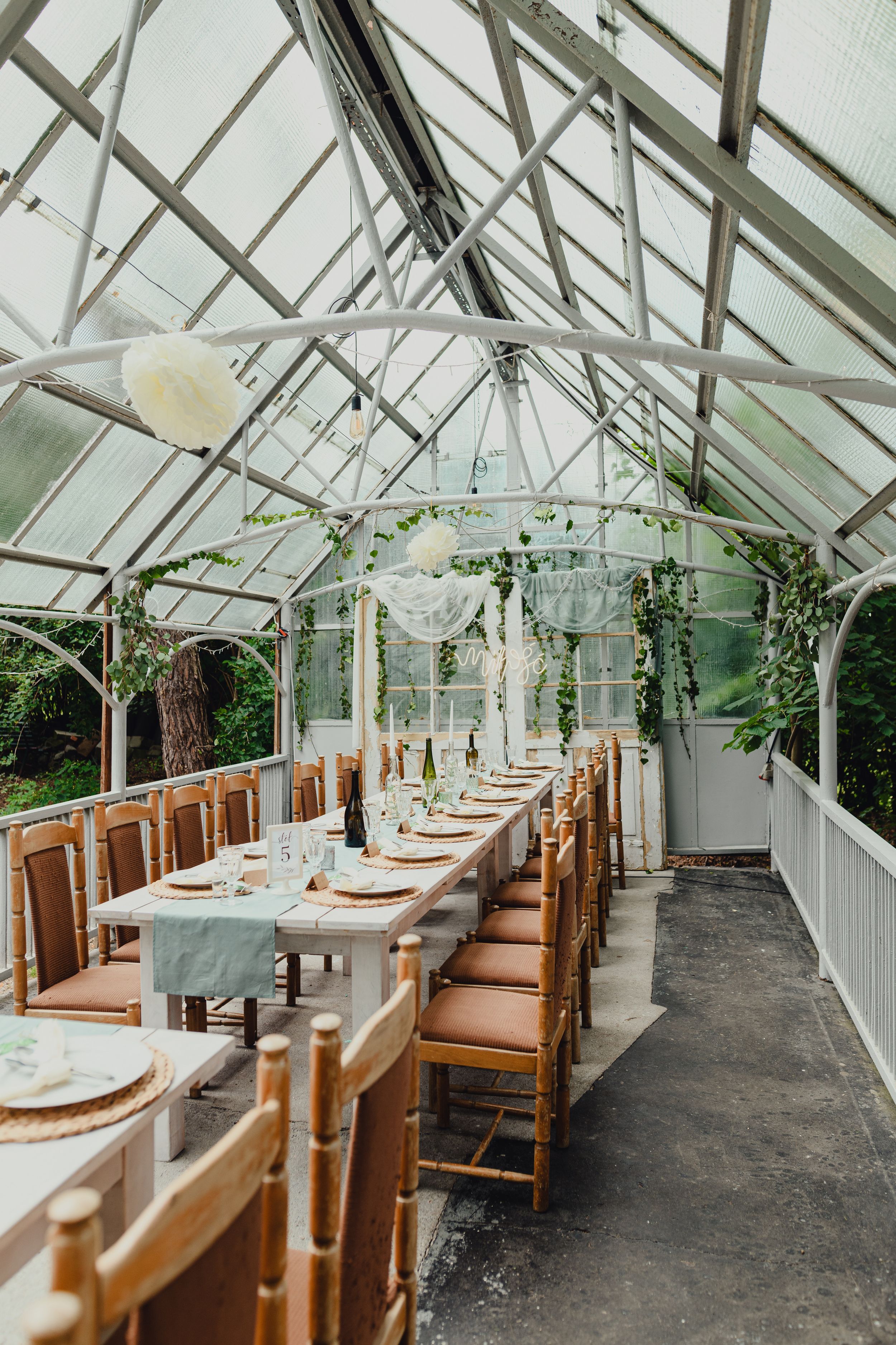 Elegant greenhouse wedding reception setup with long wooden tables and white linens under a glass ceiling.