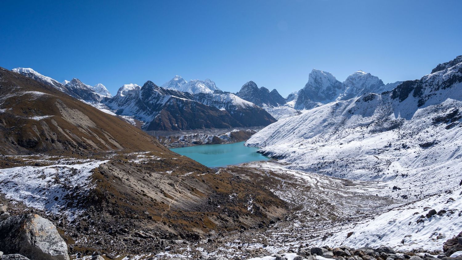 Expansive view of snow-covered Himalayan mountains with a turquoise glacial lake in the valley.