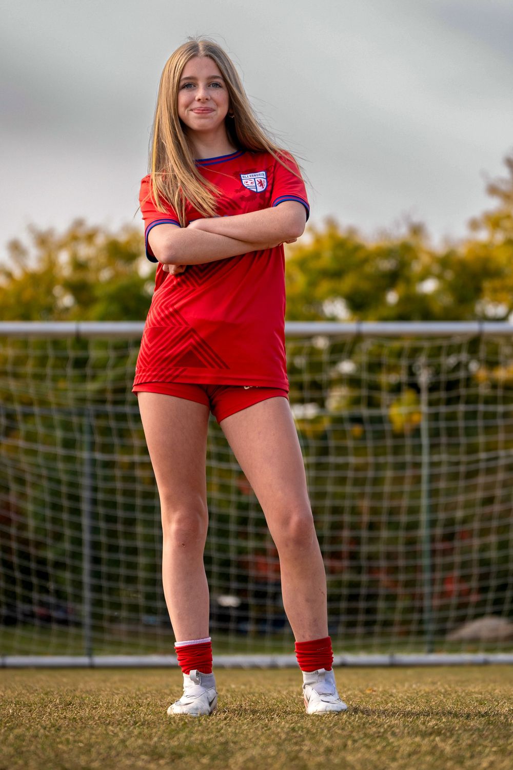 Soccer player in red uniform stands confidently in front of goal net on grass field during golden hour.