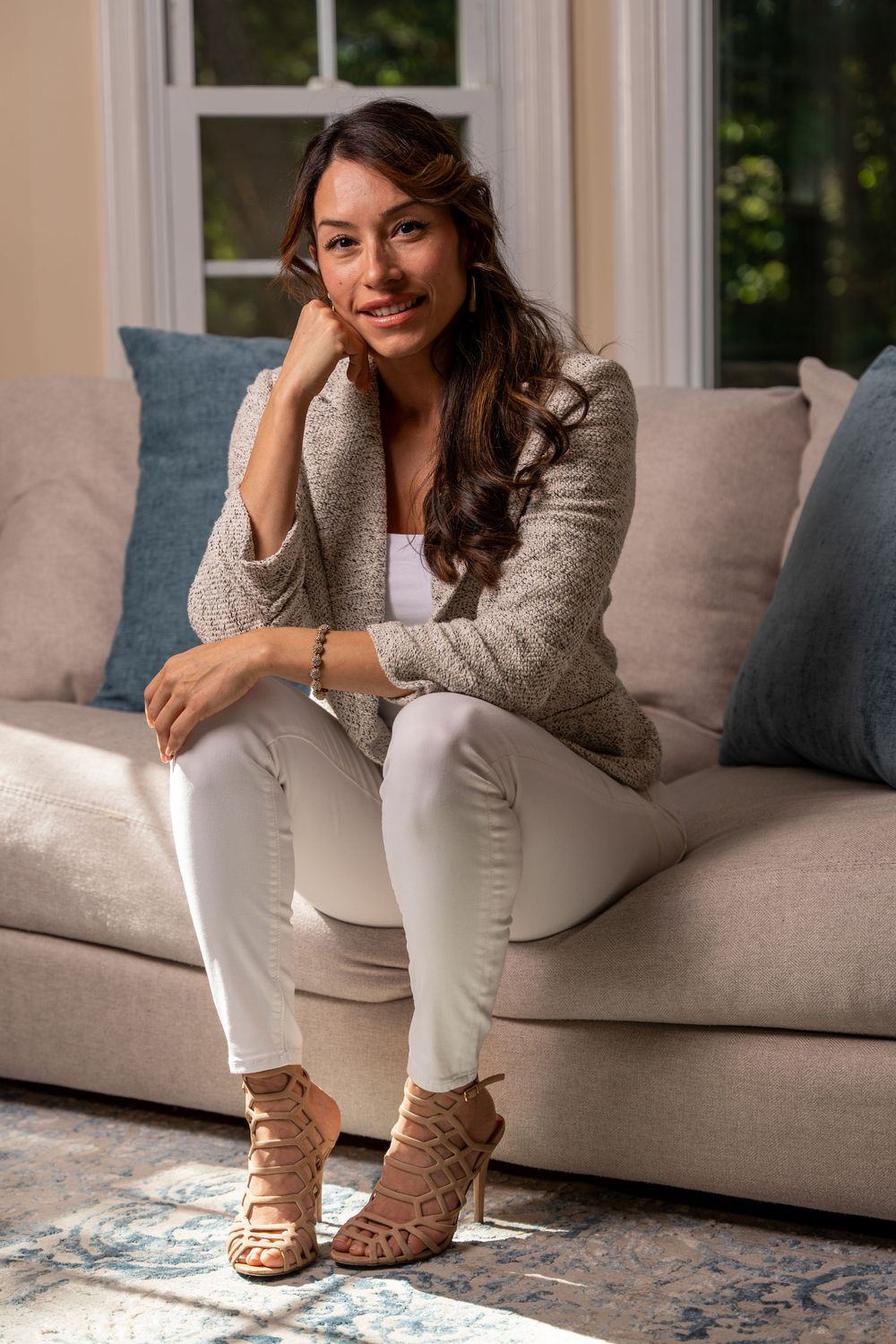 A smiling woman in white pants and a beige cardigan sits relaxed on a cream sofa with blue pillows.
