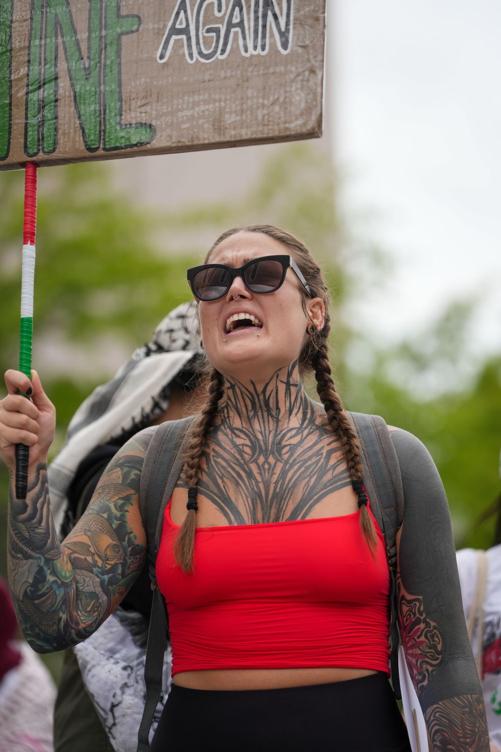 Person in sunglasses and red crop top with visible tattoos smiles while holding a sign at an outdoor event.