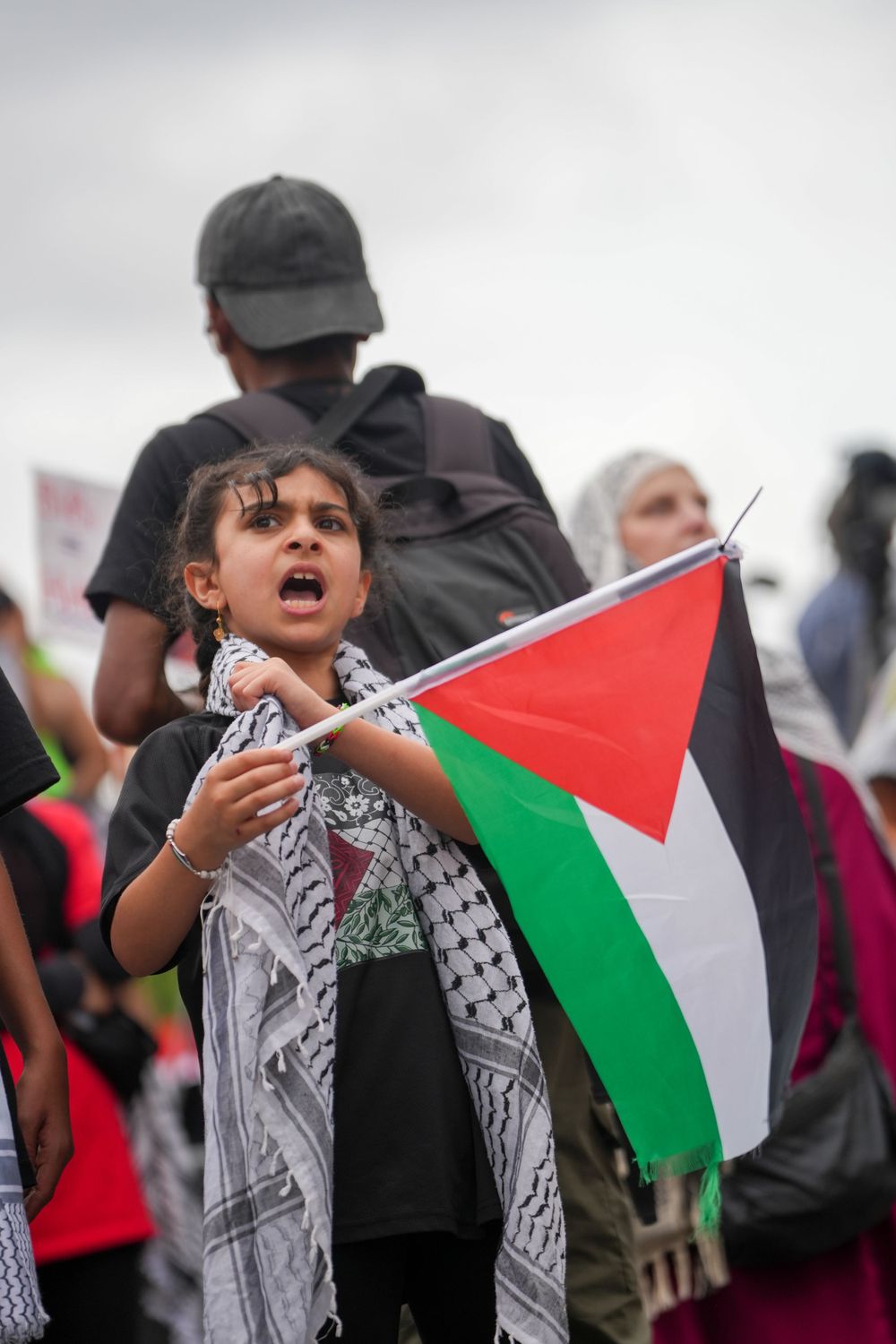 A young protestor holds up a Palestinian flag at a rally while wearing a traditional keffiyeh scarf.