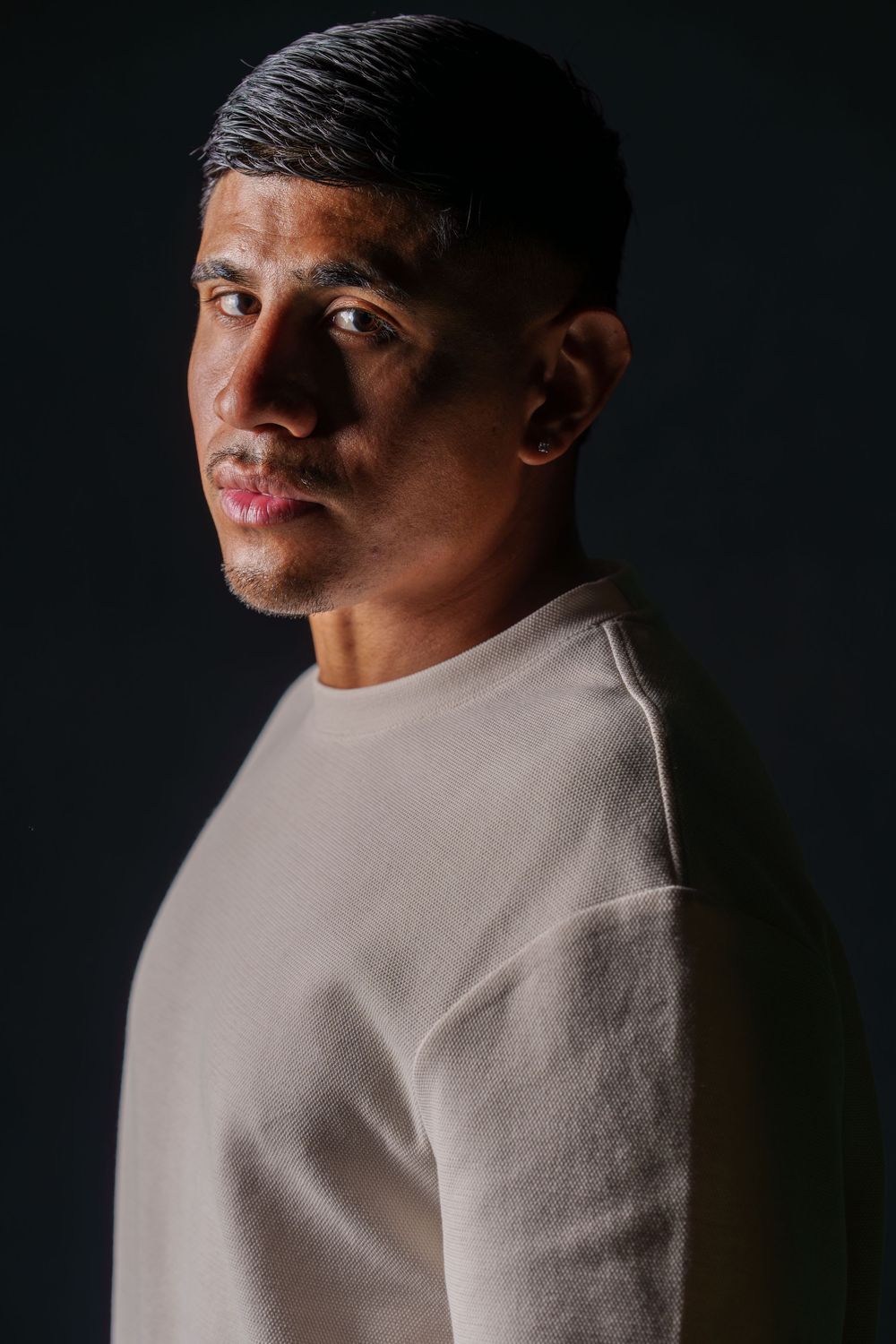 A young man in a beige sweater poses against a dark background in a dramatic studio portrait.
