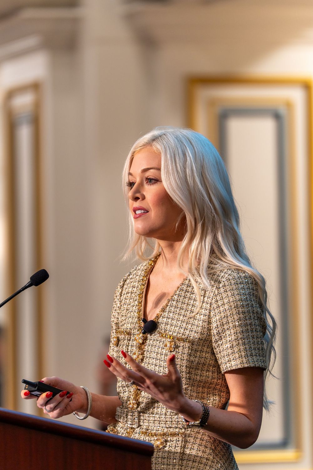 Blonde woman in gold tweed jacket speaking at podium during professional conference presentation.