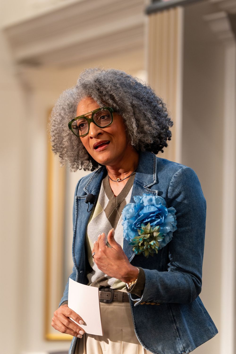 Elegant older woman with gray curly hair and green glasses holds blue flowers and papers while speaking at an event.