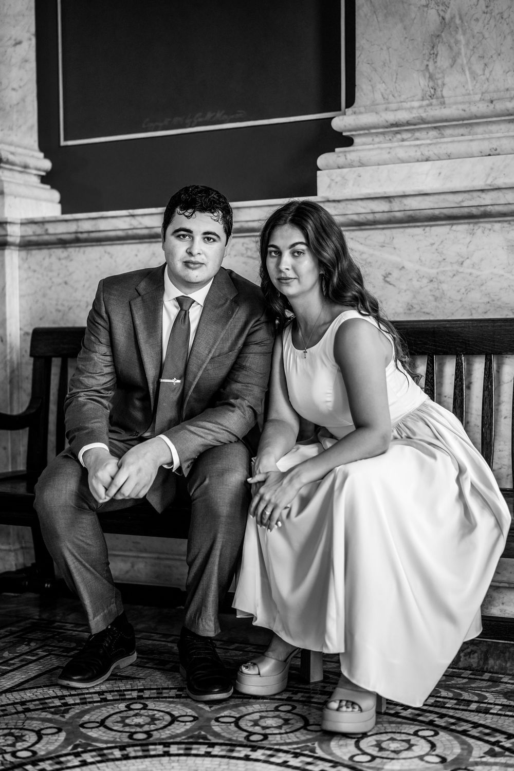 Black and white photo of a couple in formal attire sitting on a bench in an elegant marble interior.