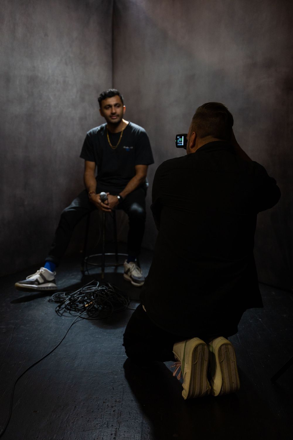 Two men in a dark studio, one seated holding a mic being filmed by another with a camera.