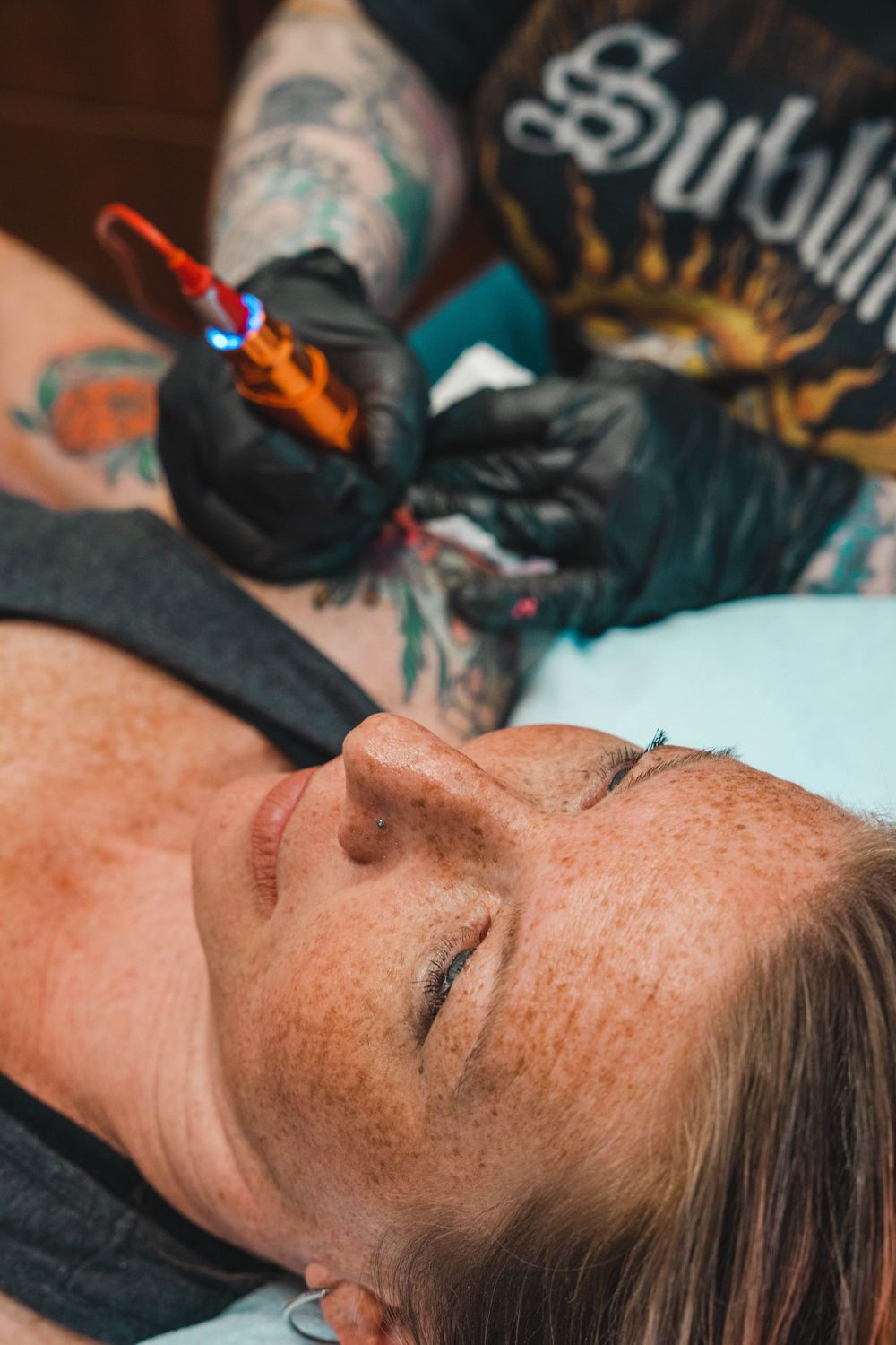 Tattoo artist in black gloves tattooing a freckled woman's face as she lies with eyes closed.