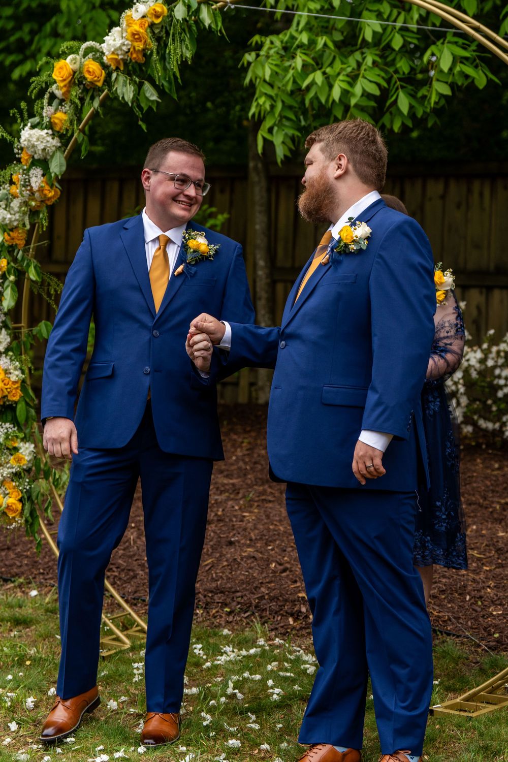 Two grooms in matching blue suits with yellow ties hold hands at an outdoor wedding ceremony.