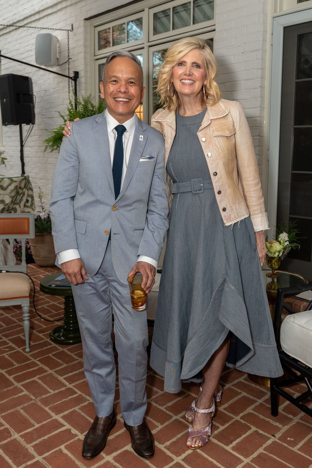 A man in a gray suit and a woman in a gray dress and beige jacket smile together on a brick patio at a social event.