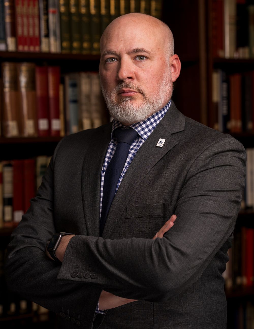 Professional portrait of a person in a suit and checkered tie standing against a library bookshelf background.