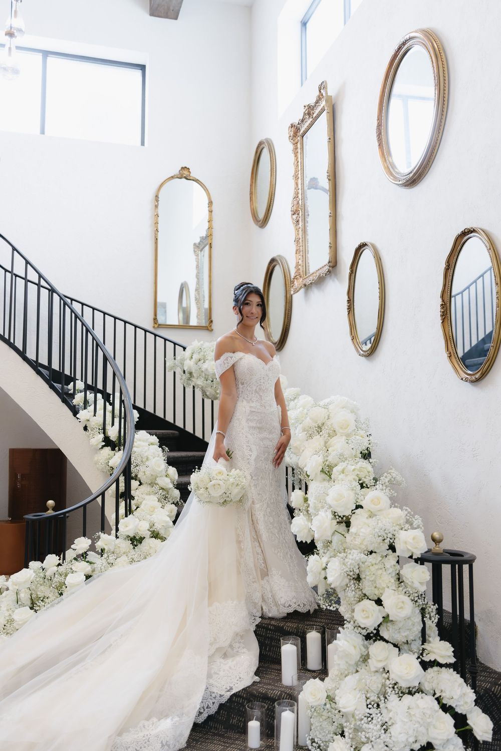 Elegant white wedding dress displayed in a bright room with ornate gold mirrors and white floral arrangements on a staircase.