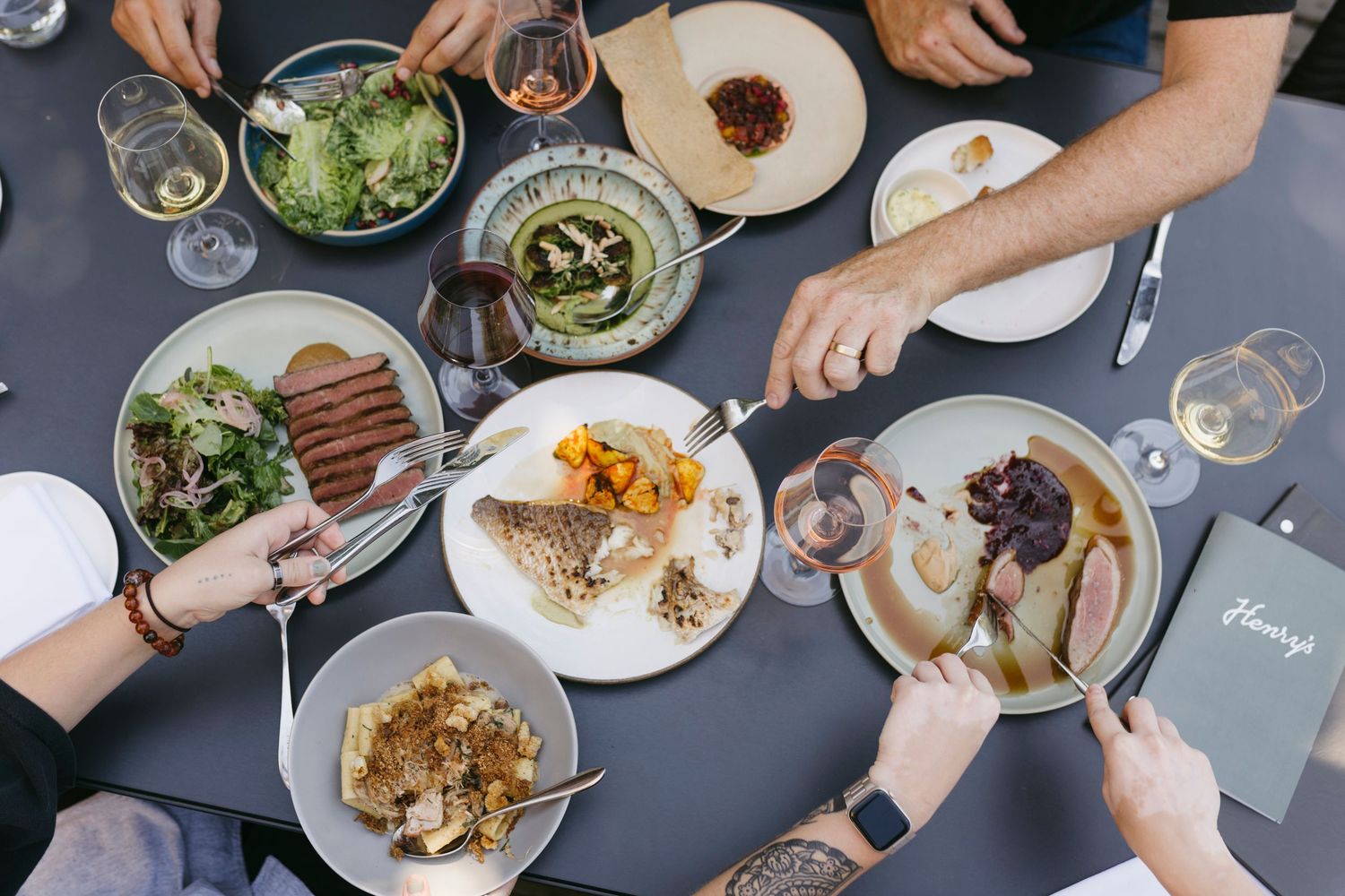 Overhead view of people sharing multiple dishes and wine glasses at a restaurant table.
