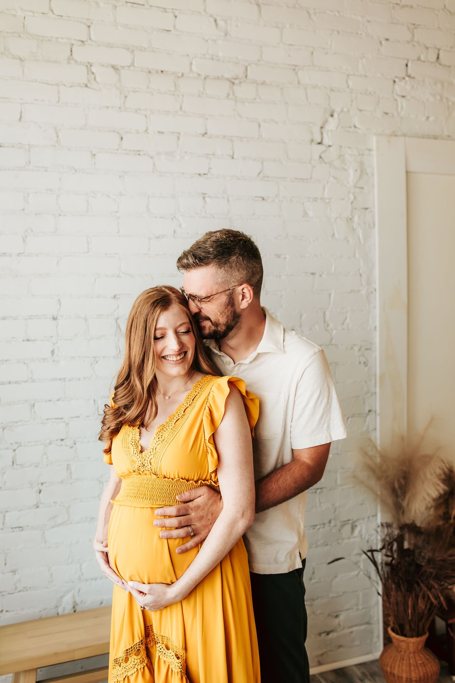 Couple shares tender embrace as they celebrate their pregnancy in a minimalist indoor setting with white brick walls.