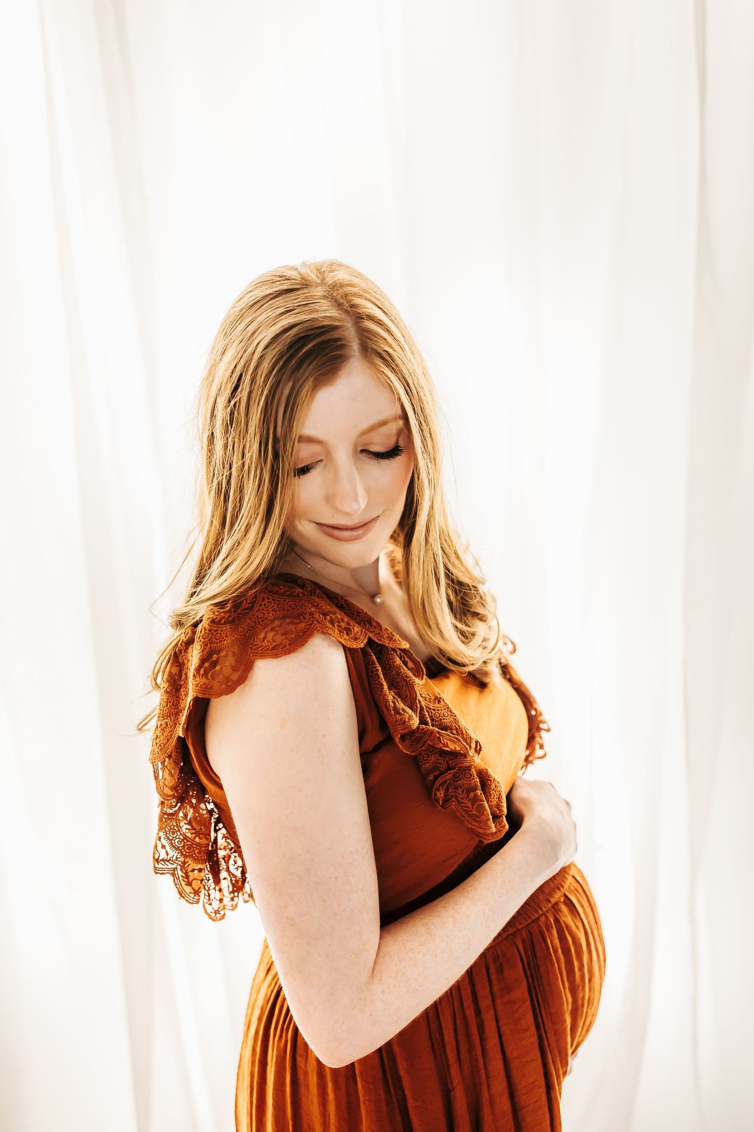 Expectant mother in rust-colored lace dress gently cradles her baby bump against white backdrop.