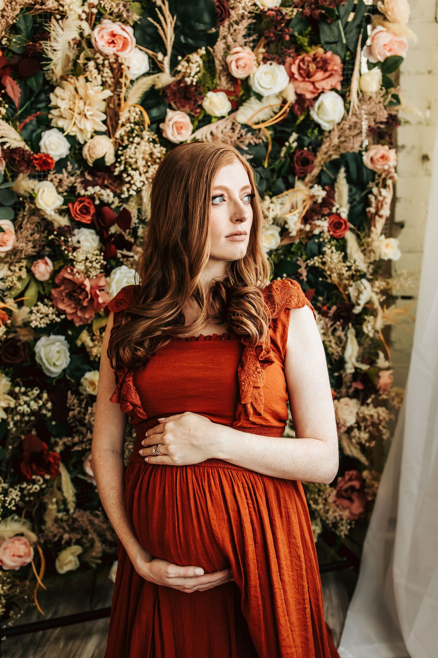 Beautiful maternity photo with long wavy hair in rust colored dress against floral backdrop of roses and peonies.
