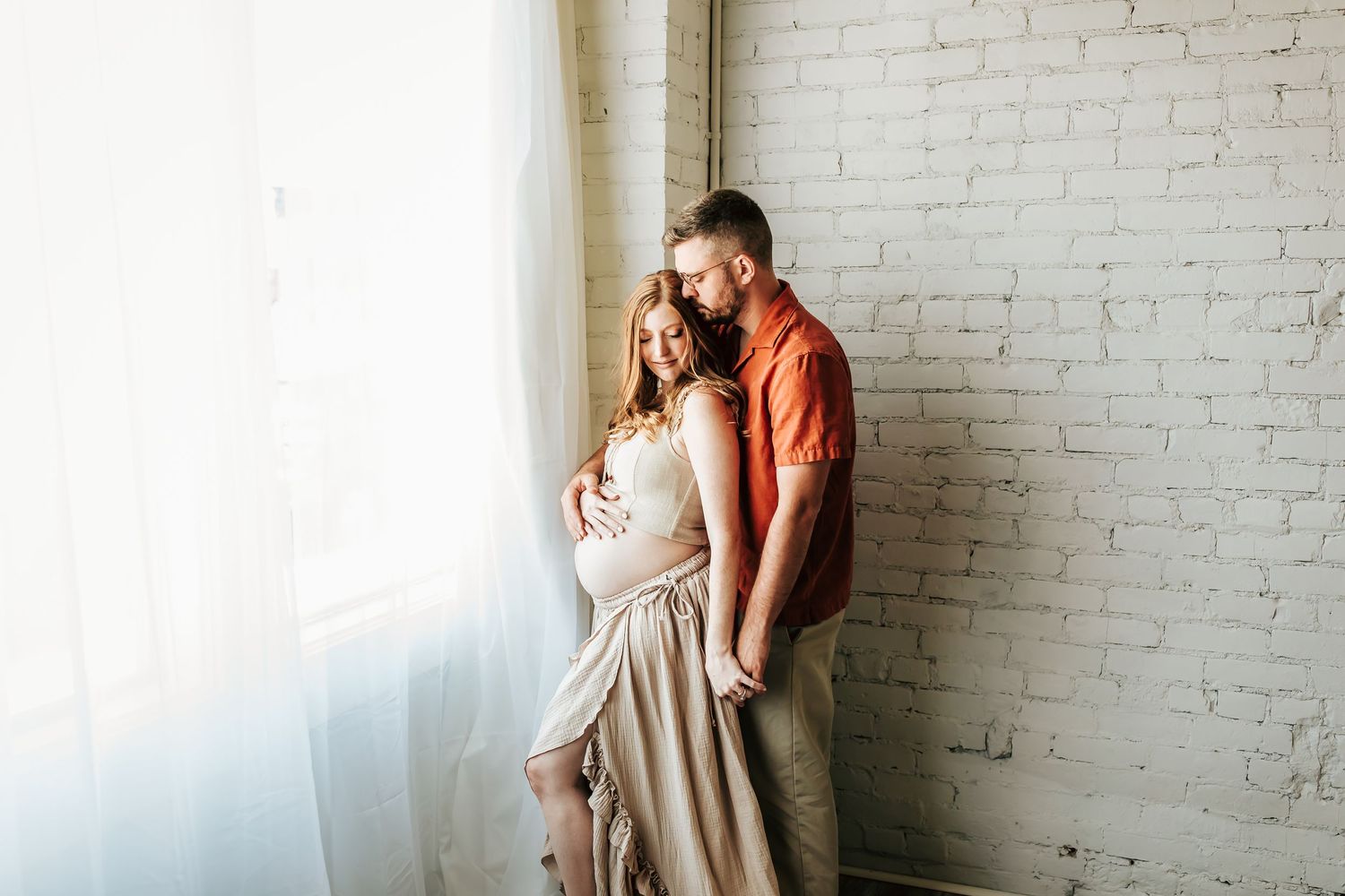 A romantic couple shares a tender moment by a sunlit window against a white brick wall while wearing coral and neutral colors.