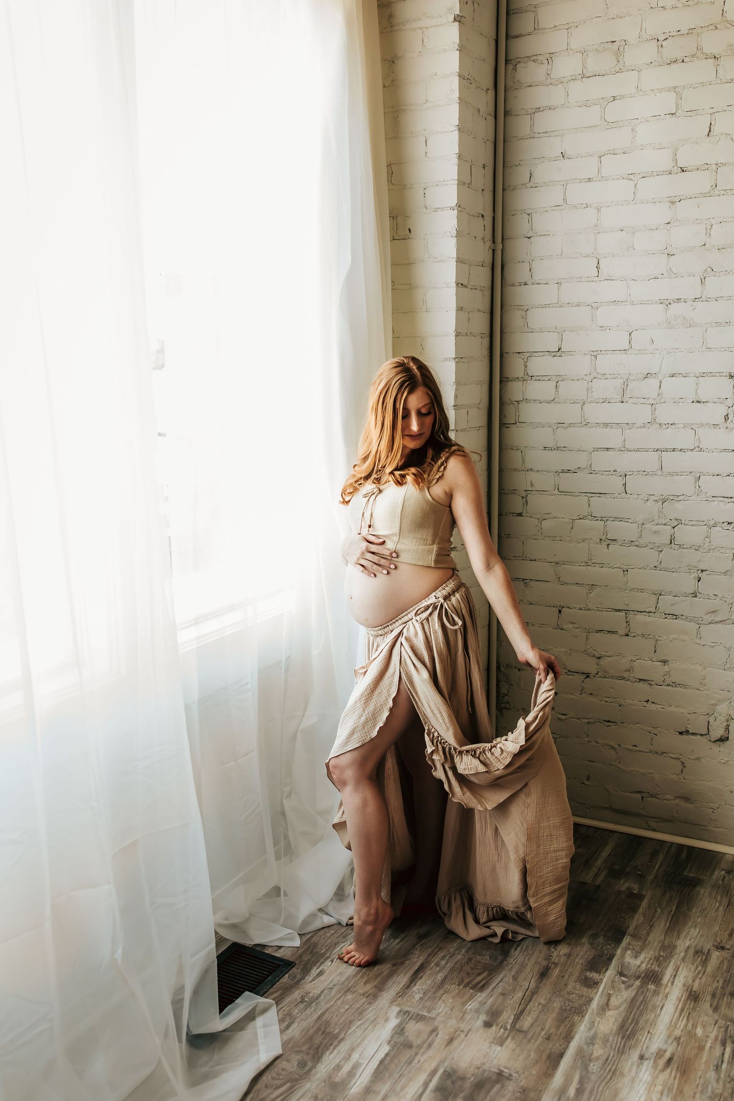 Expectant woman in beige dress poses elegantly by window with sheer curtains in rustic room with white brick wall.