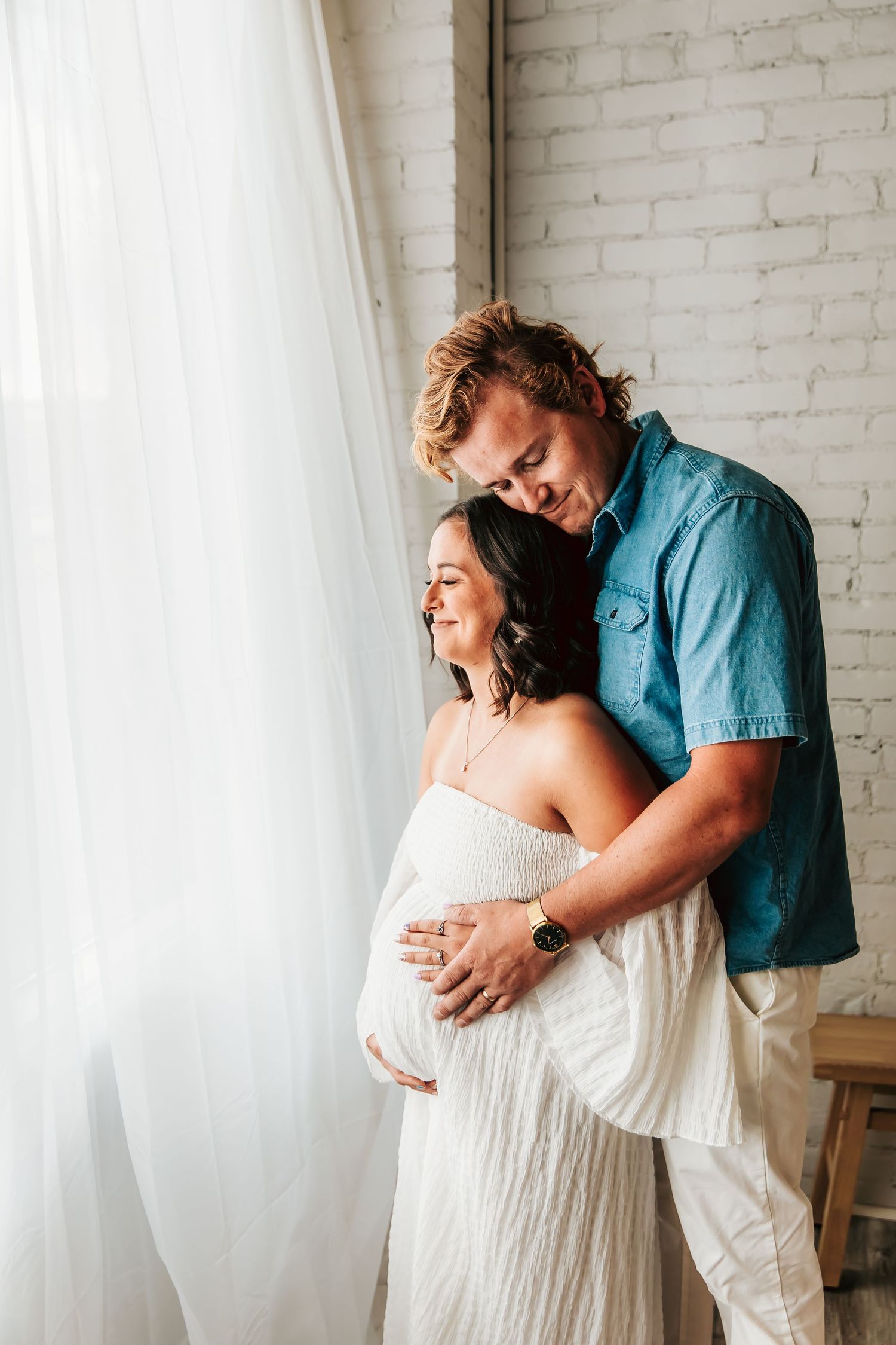 Couple embraces by window during maternity photoshoot, wearing white dress and blue shirt in front of brick wall.