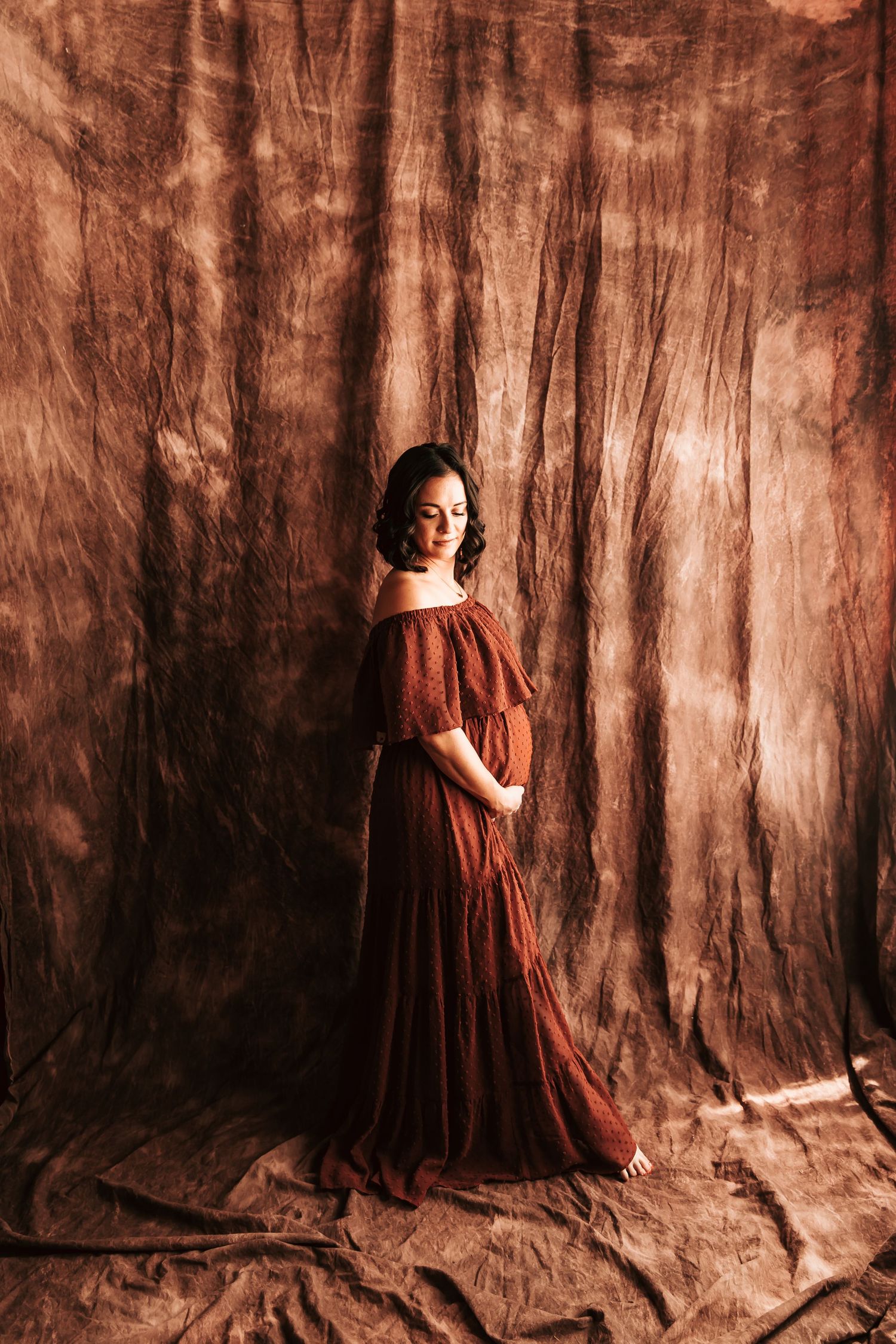 Pregnant mother in a flowing rust-colored dress poses against a textured brown backdrop in a moody studio portrait.