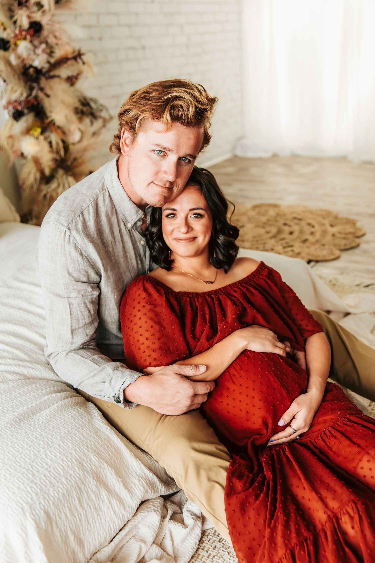 A romantic maternity photo with a couple reclining on a bed, featuring a red polka dot dress and soft natural lighting.