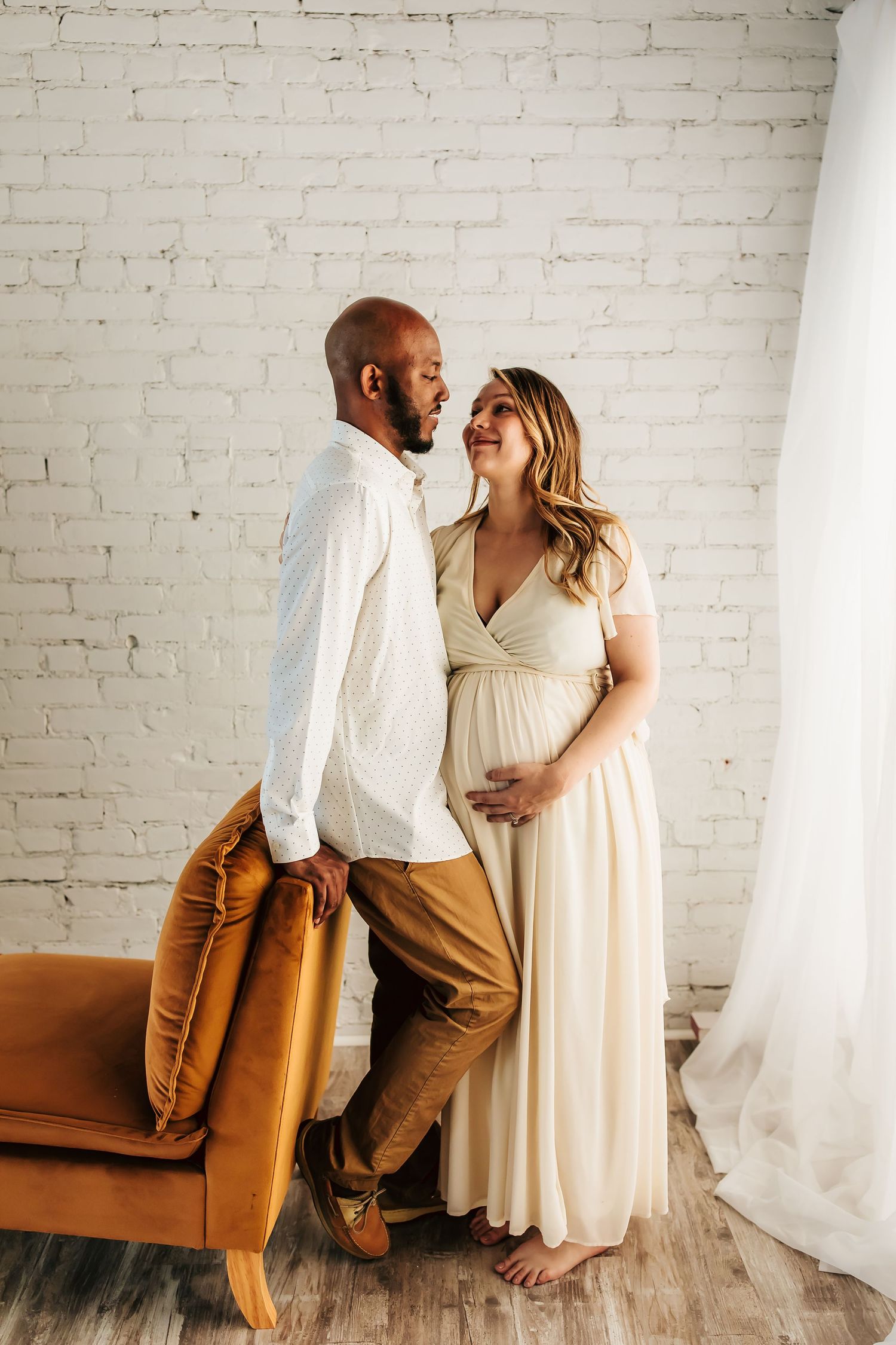 A couple shares a tender moment as they pose together near a rust colored chair against a white brick wall.