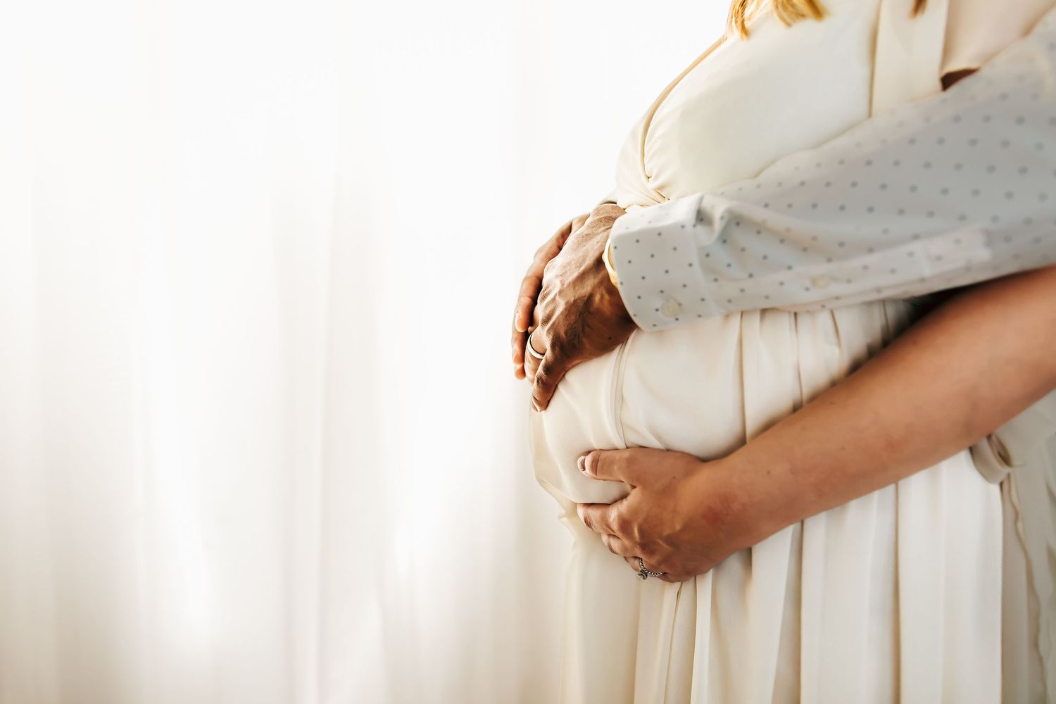 Gentle embrace captured in soft light showing a pregnant belly in a flowing white dress.
